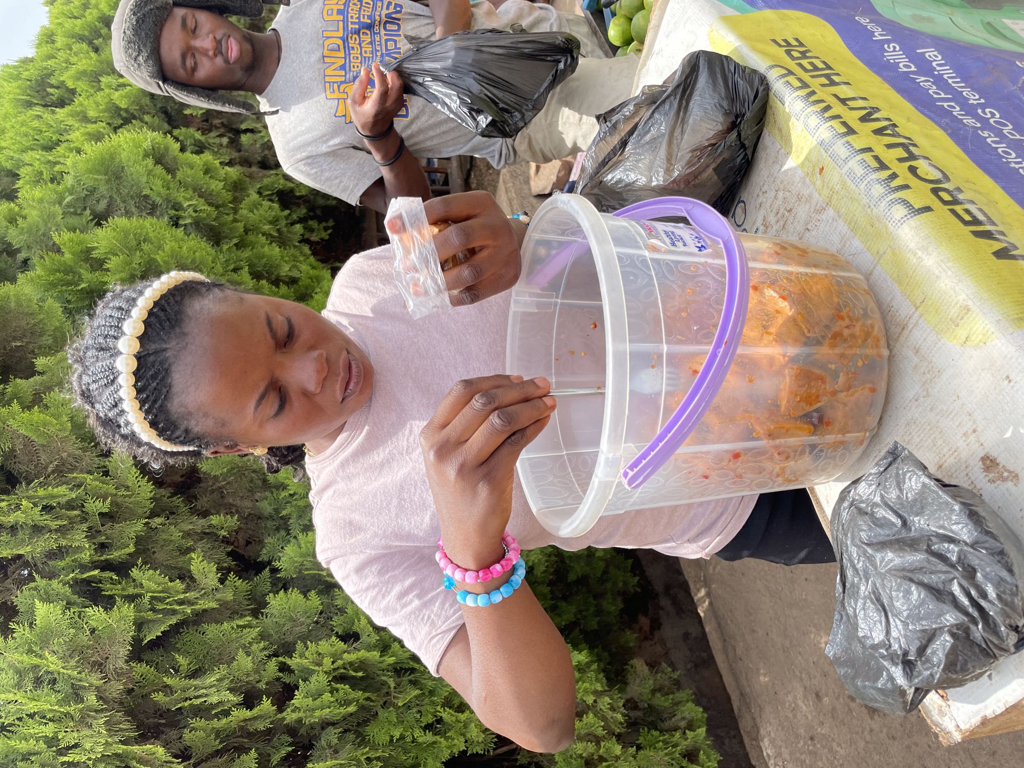 This image, captured in Jos, Nigeria, features two individuals engaged in activities at what appears to be a market stall or roadside stand under bright daylight.
In the foreground, a woman with braided hair and a pearl headband is wearing a light pink t-shirt and colorful pink and blue beaded bracelets on her right wrist. She is focused on handling small pieces of food, possibly stewed meat or fish, from a clear plastic bucket labeled "Healthy Livin Bucket." She holds a small, clear plastic bag in one hand, suggesting she is portioning or packaging the food.
Behind her, partially framed by lush green tree foliage, a man wearing a gray t-shirt and a fur-lined hat is visible. His shirt displays text including "FINDLAY BOYS TRACK AND FIELD ATHLETIC CONFERENCE CHAMPIONS." He is also seen handling black plastic bags.
The setting includes a counter or table surface where the woman's bucket rests. A prominent yellow and blue fabric banner is spread across this surface, bearing the text "PREFERRED MERCHANT HERE," "tions and pay bills here," and "POS terminal," indicating a commercial point of sale. Black plastic bags and some green produce, resembling mangoes, are also visible on the counter. The scene conveys a moment of commerce in an outdoor environment.
