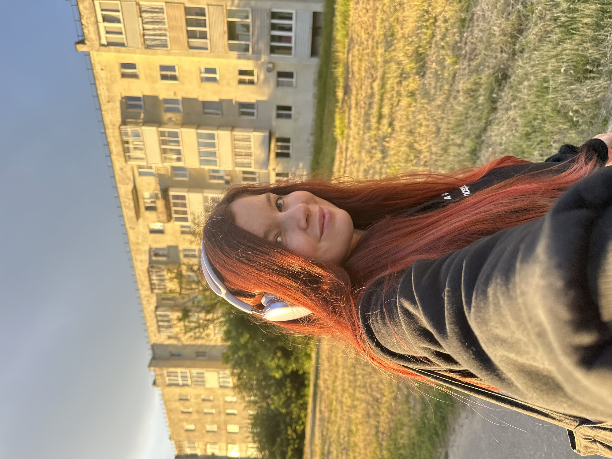 A young woman with red-tinted hair and wearing white headphones is taking a selfie. She's wearing a black hooded sweatshirt. Behind her, there's a Soviet-era apartment building with multiple windows. The light indicates it's either sunrise or sunset. In the background, there's a grassy area. Given the architecture and general ambiance, the location is likely within the city of Vladimir, Russia. The selfie seems to be taken at ground level.