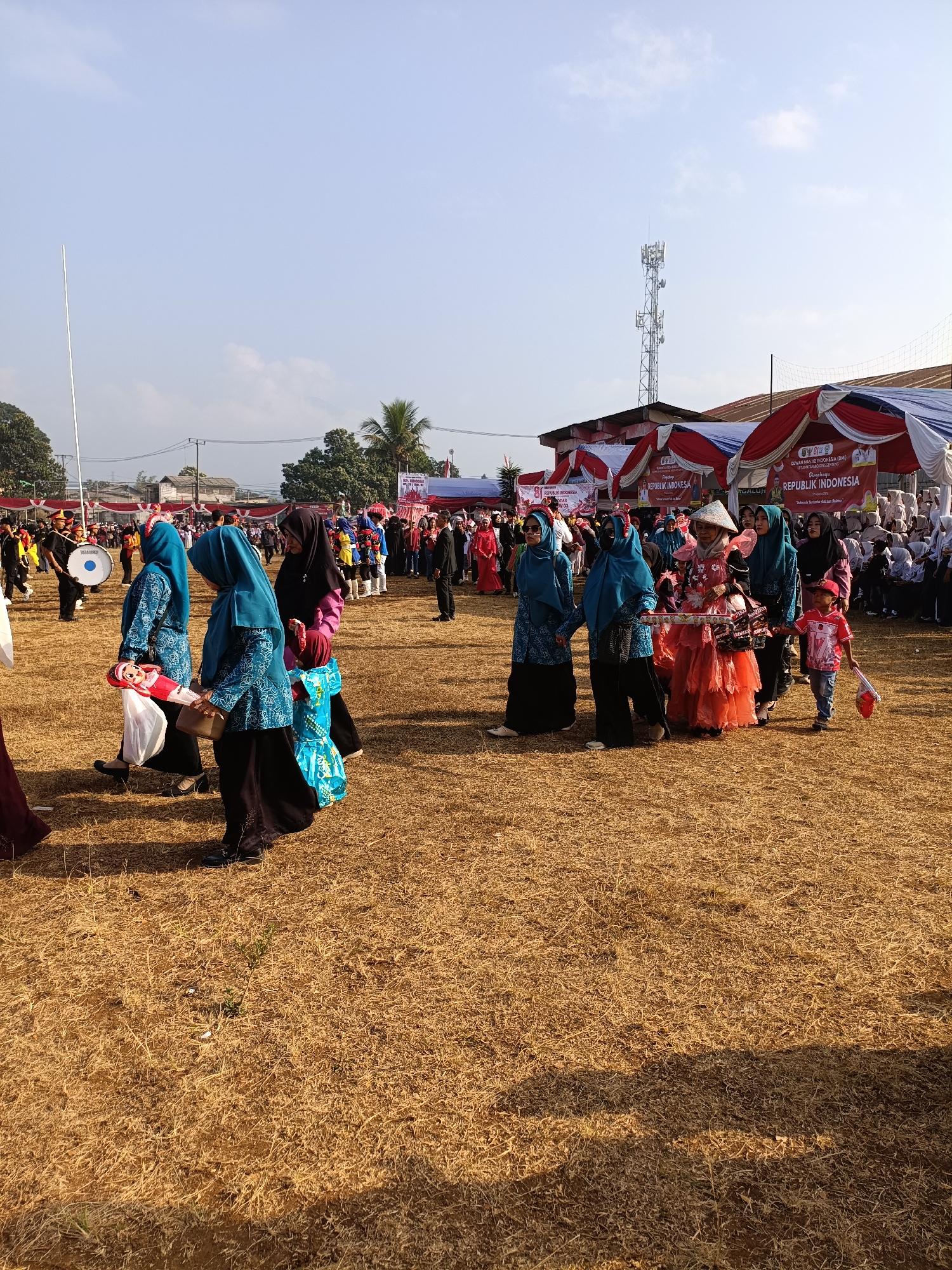 The image captures a scene in Cibodas, Indonesia, during what appears to be a parade or procession. The ground is covered in dry, yellowed grass, indicative of a warm, possibly dry season. In the background, several white and red tents are set up, suggesting an event or gathering. A prominent cell tower is visible against a clear blue sky.
The foreground features a group of people participating in the procession. Several women are dressed in colorful attire, including blue headscarves and patterned tunics, some carrying what appear to be decorative items or dolls. One woman in the center wears an elaborate orange dress and a conical hat. A young boy in a red shirt and hat is also visible, holding a small flag. Behind them, other participants are visible, including individuals in military-style uniforms, some carrying drums. Further back, a crowd of onlookers is gathered near the tents. The overall atmosphere seems festive.