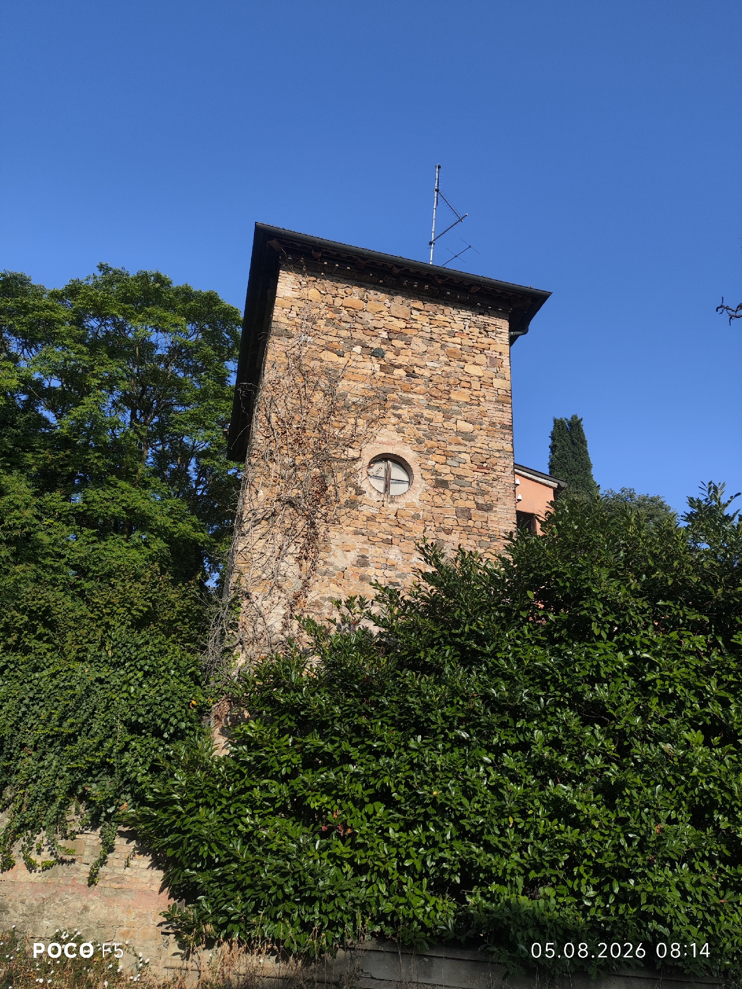A stone tower, partially obscured by lush green foliage, stands under a clear blue sky. The tower, constructed from irregular-shaped stones of varying earthy tones, features a dark, sloped roof. A small, circular window with a cross-shaped pane is visible on its facade, along with dry vines climbing its surface. An antenna array is mounted on the roof. In the background, a slender, dark green tree rises above the tower, and a section of an orange-colored building is visible. Dense green bushes and ivy cover the base of the tower and a stone wall. The lower left corner of the image displays "POCO F5" text, and the lower right corner shows the date and time "05.08.2026 08:14". The scene appears to be in Collecchio, Italy.