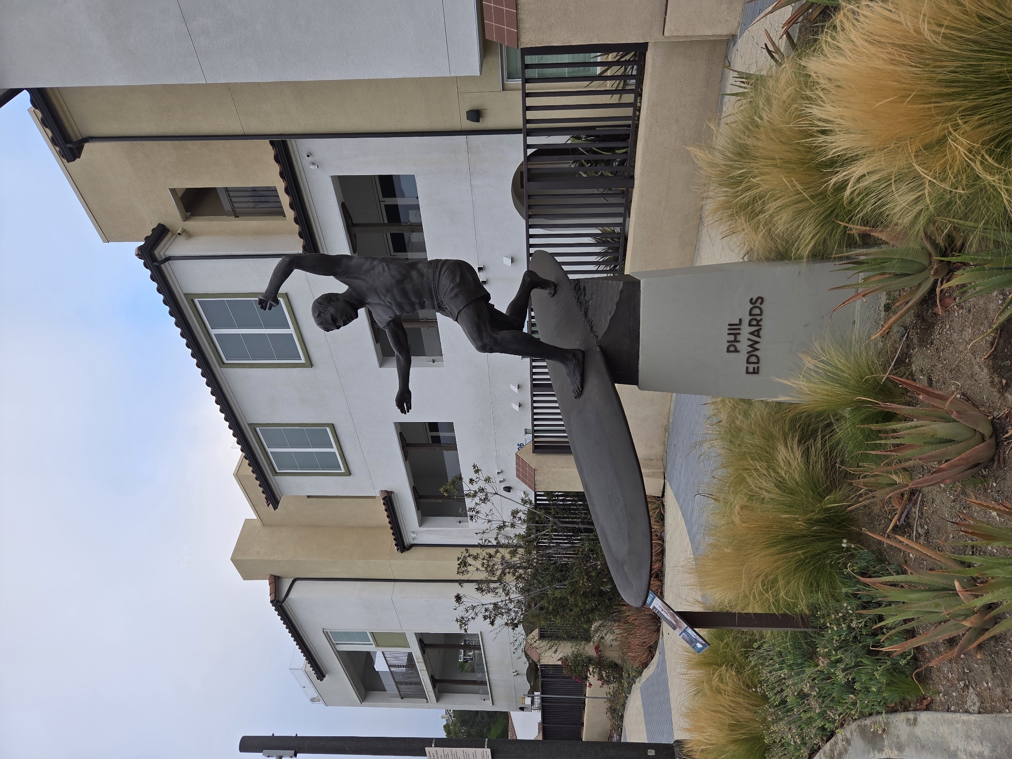 The image shows a bronze statue of a surfer named Phil Edwards in Dana Point, California, USA. The statue depicts Edwards in mid-ride on a surfboard, with one arm raised. The sculpture stands on a concrete pedestal inscribed with "PHIL EDWARDS." It is situated in front of a modern building with a Mediterranean architectural style, featuring stucco exteriors, red tile roofs, and balconies. The surrounding landscaping includes drought-tolerant plants such as ornamental grasses and succulents. A paved pathway with a diamond pattern is visible in the foreground, leading towards the statue. The sky is overcast.