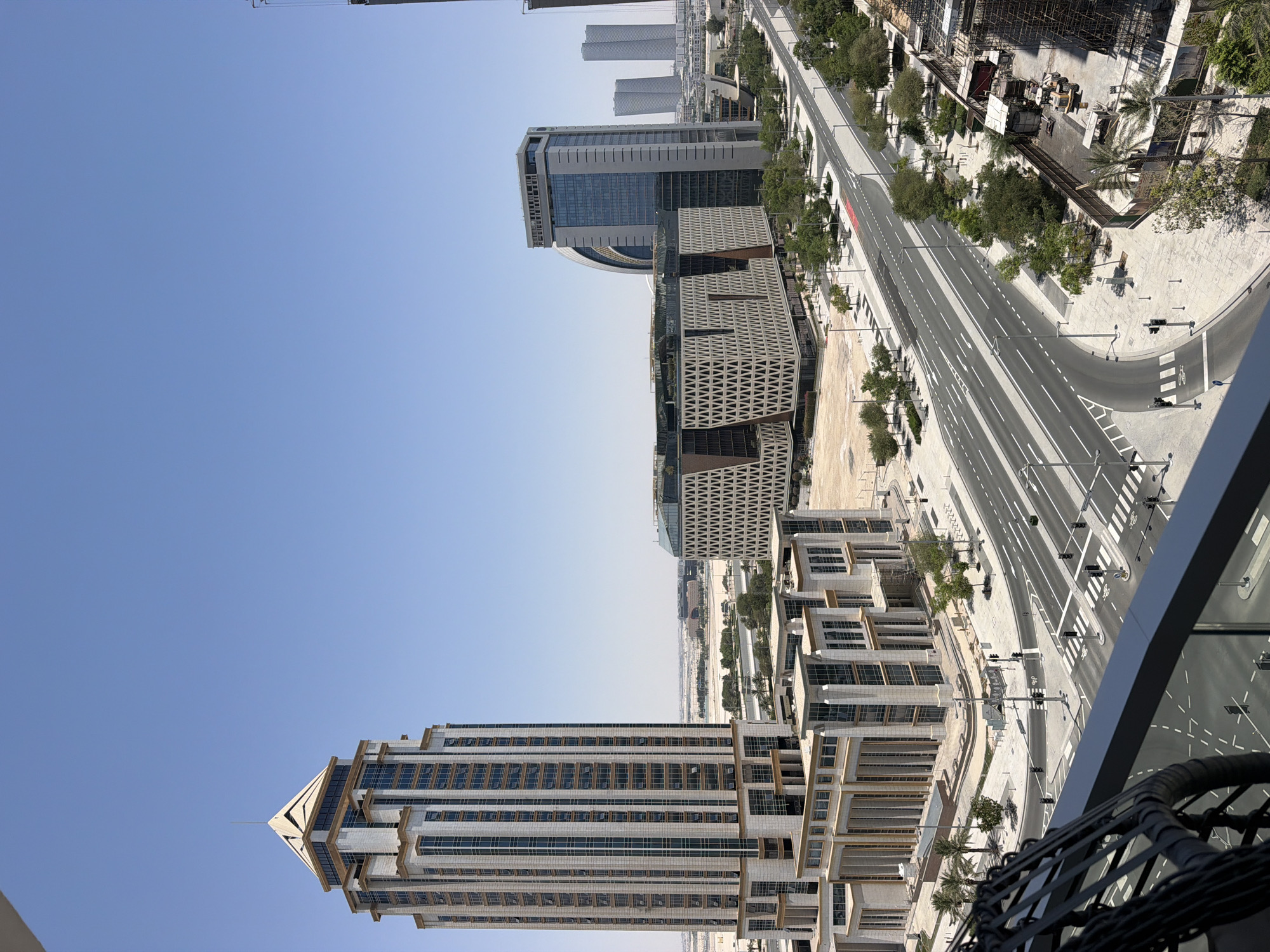 This media file shows an aerial view of a modern urban landscape under a clear blue sky. The scene is dominated by tall, contemporary buildings with glass facades and geometric designs. A wide, multi-lane road curves through the cityscape, bordered by trees and pedestrian walkways. Construction sites with scaffolding and heavy machinery are visible, indicating ongoing development. The perspective suggests the photo was taken from a high vantage point, possibly a balcony or another tall building. The time of day appears to be daytime, with bright sunlight casting shadows. The location is likely a city known for its modern architecture and urban planning, possibly in a desert climate given the sparse vegetation and arid-looking ground in some areas. No people are clearly visible. The overall impression is one of a thriving, developing metropolitan area.