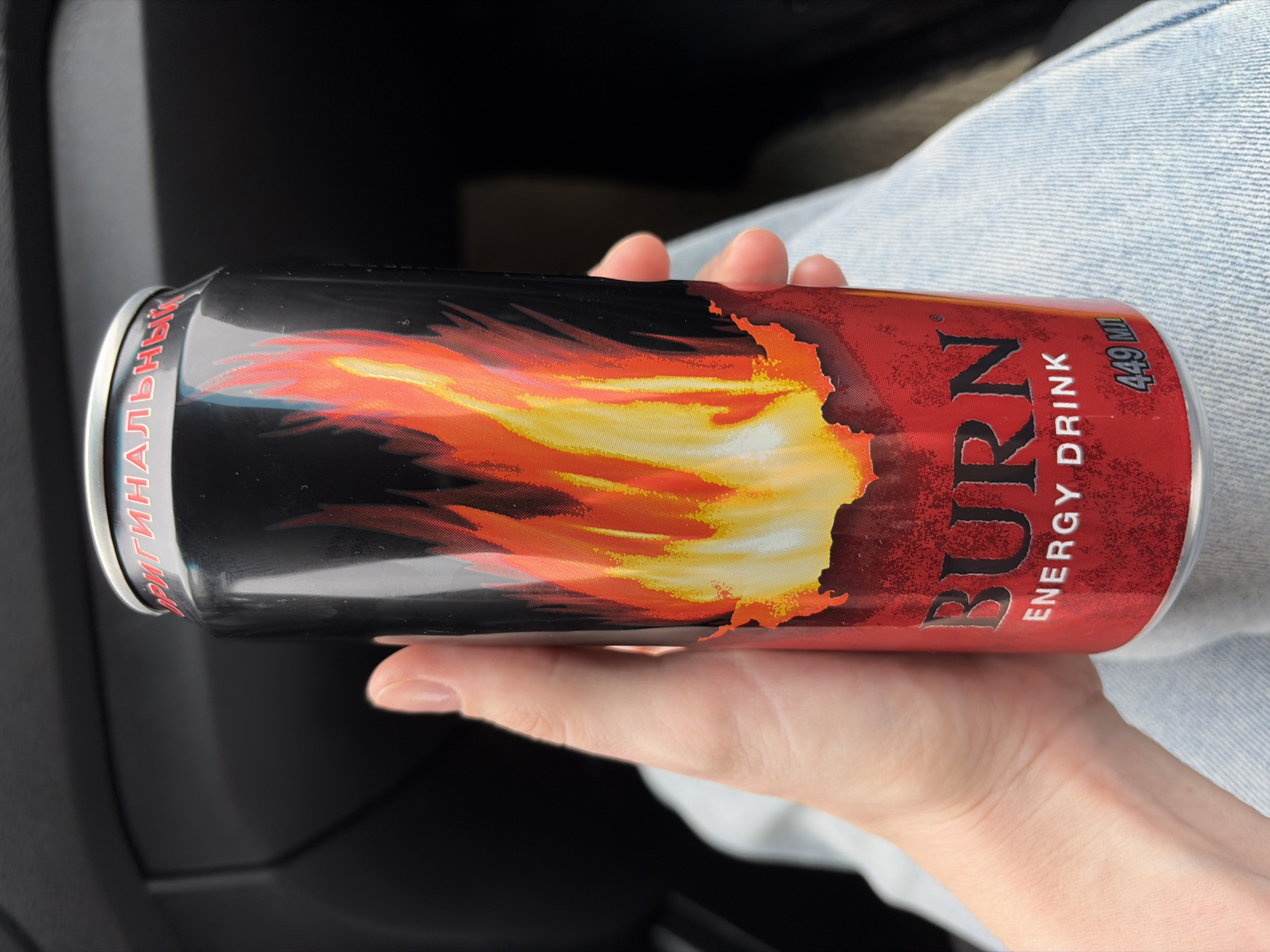 A hand holds a 449 ml can of Burn energy drink in what appears to be a car. The can is predominantly black and red with a stylized flame design. The text on the can reads "BURN ENERGY DRINK" with "449 ML" visible near the top. Part of the word "ОРИГИНАЛЬНЫЙ" is visible near the top of the can. The hand is fair-skinned, and the background includes denim fabric and dark car interior elements. The city of Прикамский, Russia is not discernible in the image.