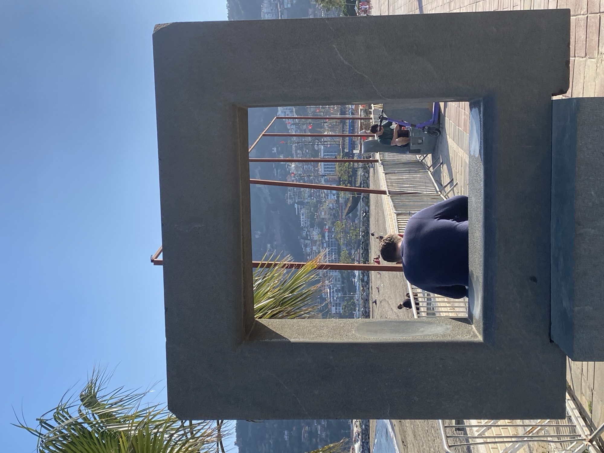 The image displays an outdoor panoramic view, framed by a prominent, dark grey, rectangular stone or concrete structure in the foreground. This frame acts as a visual aperture, through which a coastal city unfolds.
In the immediate foreground, on the right side of the frame, a person wearing a dark blue top is seen from behind, leaning into or observing the view through the frame. Further back and to the left, another individual in a green shirt is seated on a low wall or bench, with a purple scooter resting nearby. Other people are visible as smaller figures in the middle distance, suggesting a public area or promenade.
Beyond the frame, a densely built city sprawls across a hillside, with numerous buildings under a bright, clear blue sky. At the bottom of the framed view, a body of water, likely the sea, meets a coastline or beach. Lush green palm fronds are visible on the left side of the image, both outside and slightly inside the frame, indicating a warm climate.
The scene is illuminated by bright daylight, under a sunny and cloudless sky, suggesting favorable weather conditions. The overall impression is that of a popular scenic overlook, capturing a vibrant urban landscape against a natural backdrop. No visible text is present in the image. The location is Alanya, Turkey.