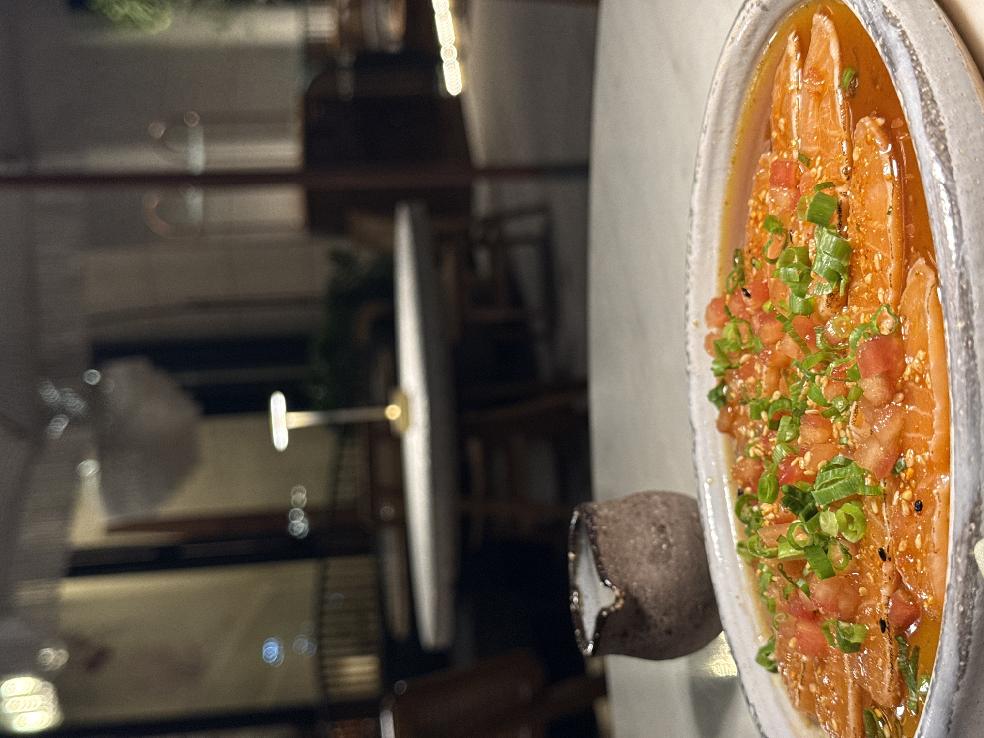 The image shows a plate of sashimi with chopped tomatoes and green onions on a white table. Next to the plate is a small, brown ceramic container with a white rim. In the background, out of focus, is an interior scene with tables and chairs, presumably a restaurant or dining area. The location is دبي, United Arab Emirates.
