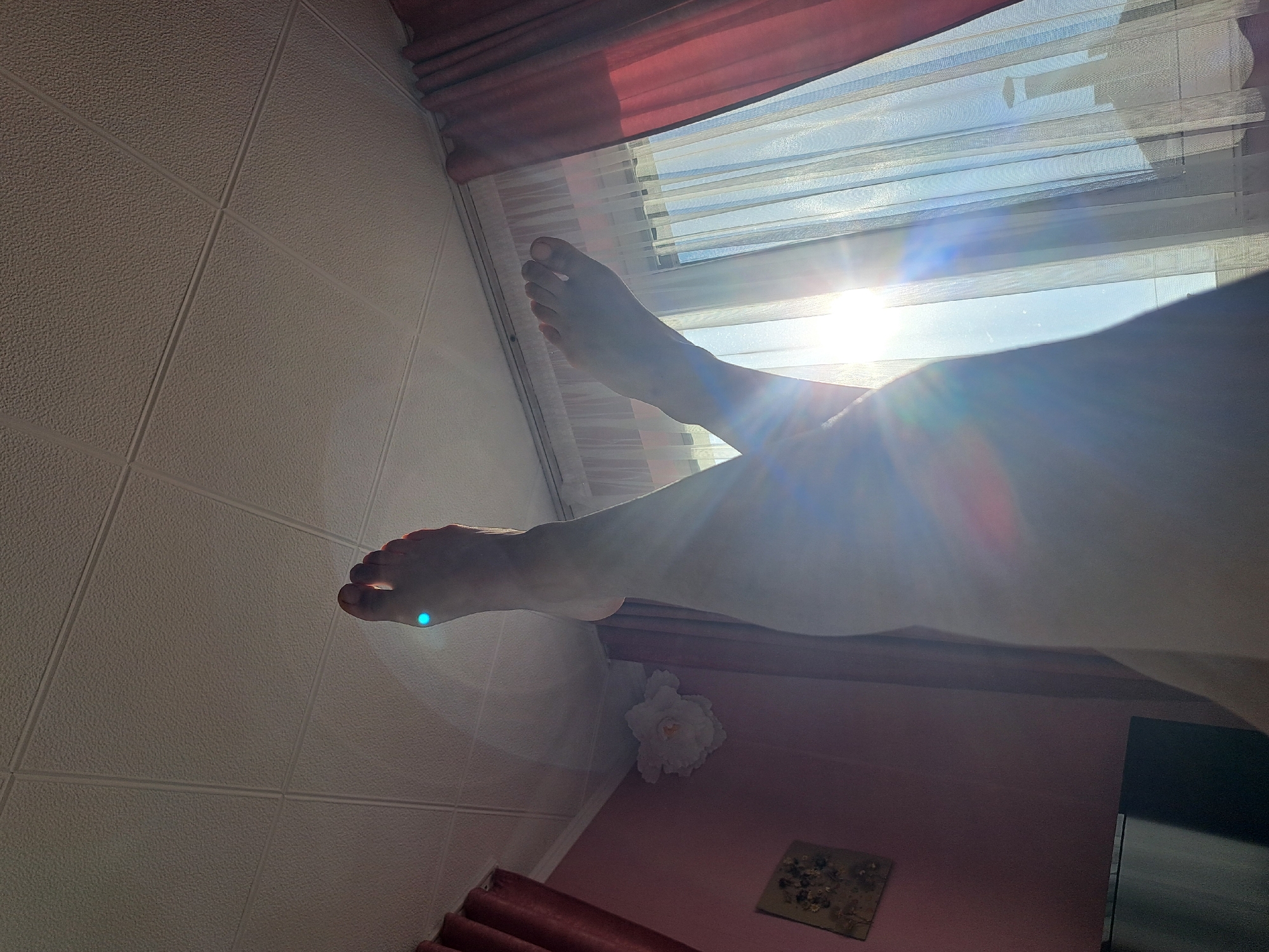 The image shows a pair of feet in front of a window. The room is brightly lit by sunlight, which washes out some of the details. The feet are crossed, with one foot closer to the window. There is a white flower decoration on the wall beneath the window. The wall is pink, and there is a small picture on it. A television is visible on the right. The window has red drapes at the top. The tiles on the wall are white and square. The location is not known.