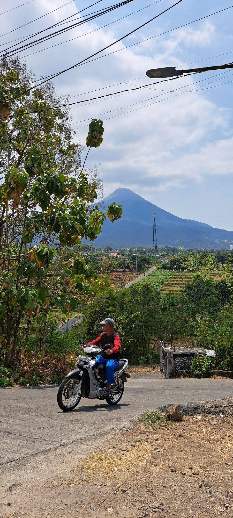 An outdoor, full-frame, vertical, eye-level shot captures a man riding a motorcycle on a paved road in Sumber Gedang, Indonesia. The man, wearing a red long-sleeved shirt, blue pants, and a grey cap, sits on a silver and black motorcycle with an Indonesian license plate. He is looking to his right. The road curves slightly uphill to the right. To the left of the road, lush green trees with some dry leaves are prominent, with a few branches extending into the foreground. Beyond the trees and the road, a village with houses and fields is visible, leading to a large, blue, conical mountain in the distance under a bright, cloudy sky. A tall cell tower is situated on the slope of the mountain. Overhead, power lines crisscross the sky, and a street light is visible on the right. The ground on the right side of the road is dry and rocky.