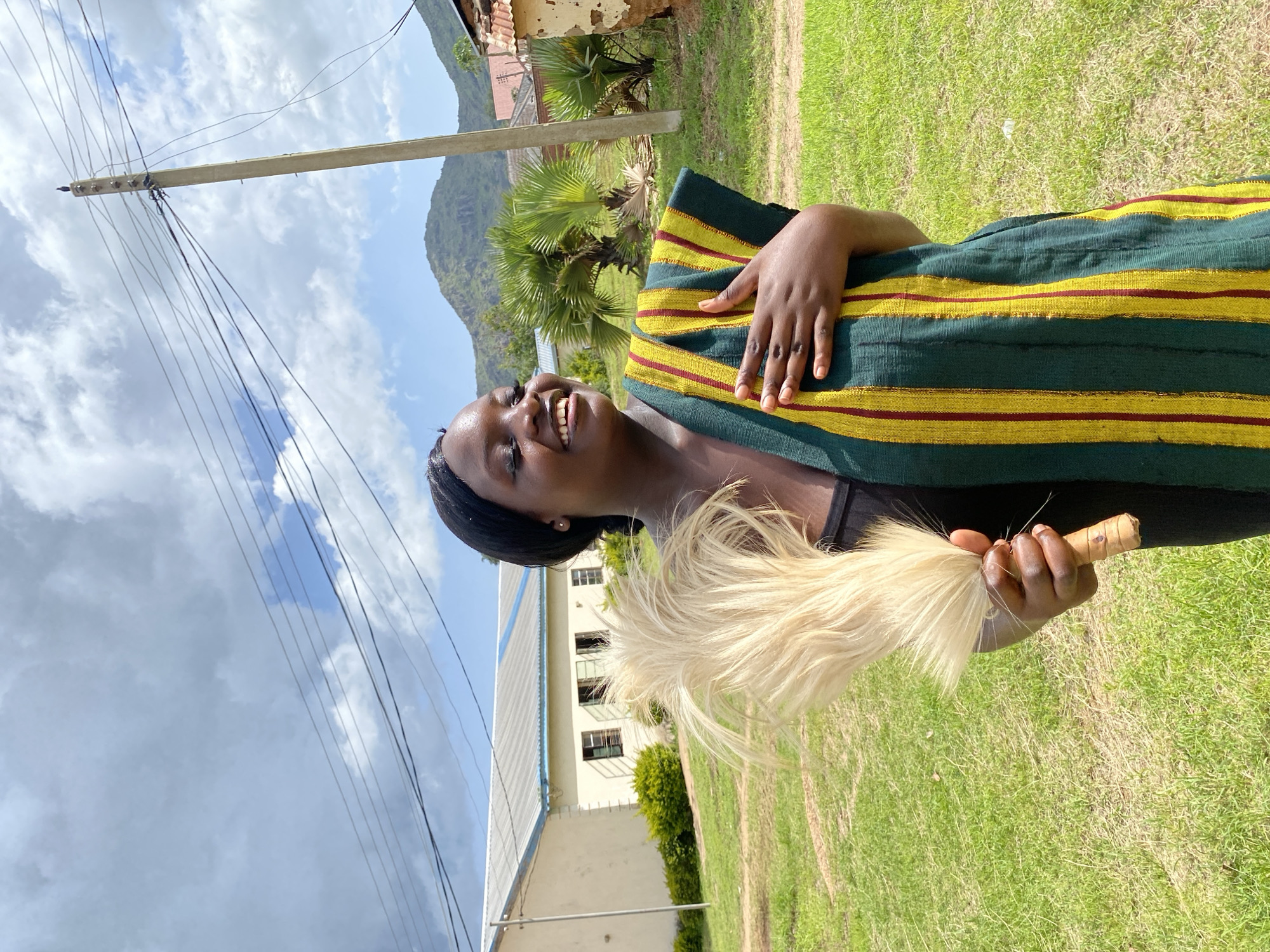 This is a portrait of a young Black woman outdoors. She is wearing a traditional garment with thick horizontal stripes of dark green, yellow, and thin red. Her left hand rests on her chest, covering a portion of the garment, and her right hand holds a light-colored whisk or fly-swatter made of animal hair. The woman is smiling broadly, with her head tilted slightly back and her eyes looking upwards. She has dark braided hair pulled back.
The setting appears to be rural or semi-rural. Behind her, there is a well-maintained grassy area. In the mid-ground, a two-story building with a white exterior and a light-colored roof is visible, along with some green shrubbery. Further in the background, there are palm trees and a lush, green mountainside under a cloudy sky. A utility pole with numerous wires extends from the left side of the frame towards the upper left corner.
The lighting suggests it is daytime, likely afternoon, given the bright sunlight illuminating the grass and the building. The weather appears partly cloudy, with patches of blue sky visible between the clouds. The overall mood of the image is joyful and relaxed, primarily conveyed by the woman's happy expression. The location context is Wang, Nigeria. There is no visible text in the image.