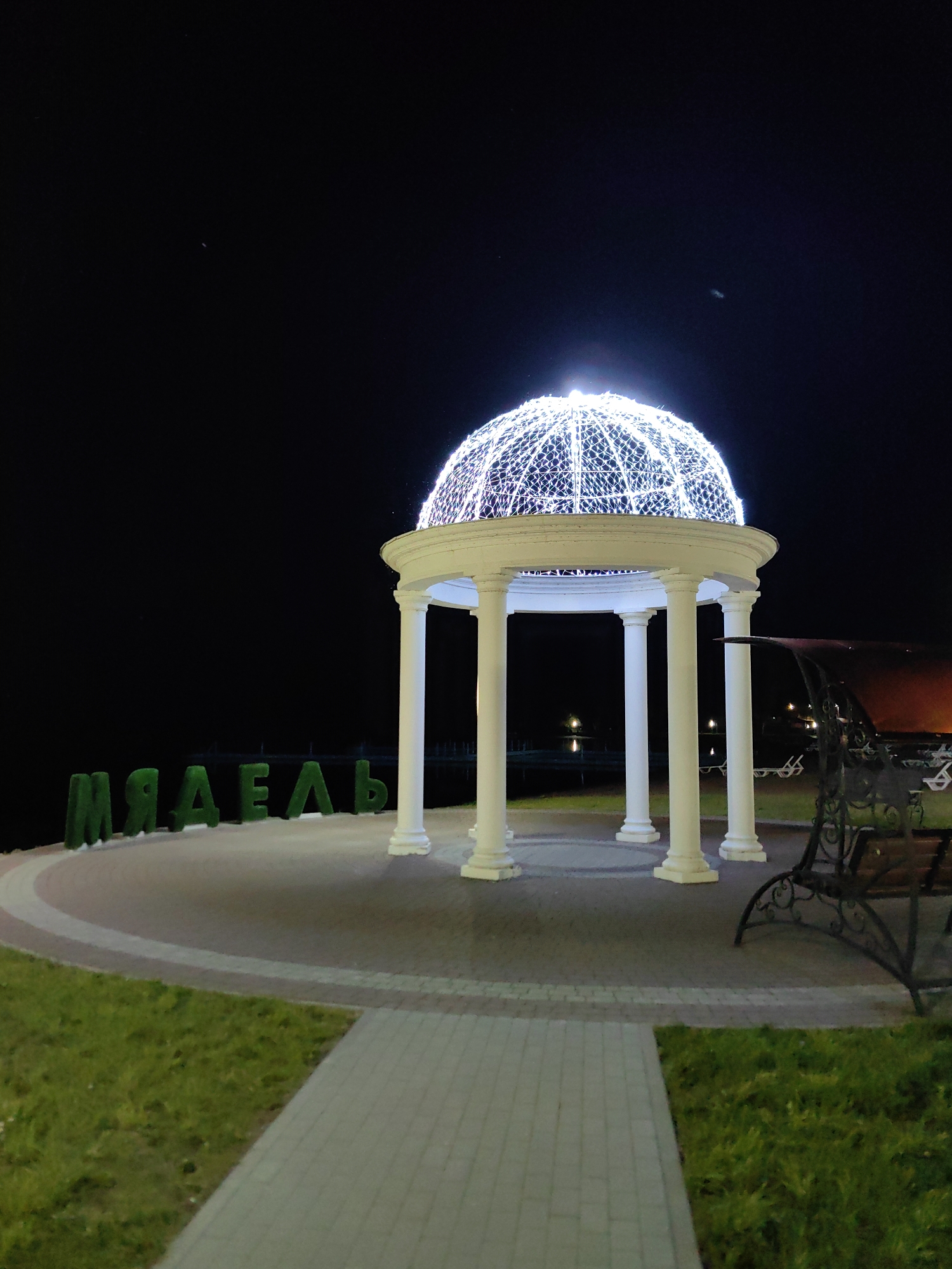 The image depicts a nighttime scene at the Myadzyel embankment in Myadzyel, Belarus. A white, domed gazebo, illuminated by white string lights, stands as the central focus. The gazebo is supported by four thick white columns. In the foreground, large green letters spell out "МЯДЗЕЧ" in Cyrillic, likely signifying the name of the town. The ground is paved with light-colored bricks, bordered by a curved section of darker paving stones and green grass. A park bench with an ornate black metal frame and wooden slats is visible to the right of the gazebo. In the background, the dark expanse of water, possibly a lake or river, is visible, with faint lights in the distance. The sky is dark and clear, with a few small, bright specks that could be stars or distant lights. The overall impression is of a well-lit public space designed for evening relaxation and tourism.
