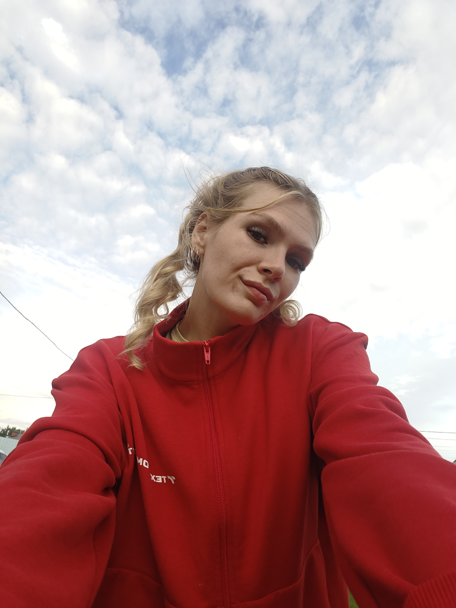 A young woman with blonde hair in pigtails, wearing a red zip-up jacket, looks up at the camera. She has on makeup, including eyeliner and lipstick. The background is a cloudy sky. The scene appears to be outdoors, possibly in a rural or suburban setting, given the presence of a telephone wire and some distant structures. No specific landmarks identifiable as being in the city of Русский Вожой, Russia are visible.