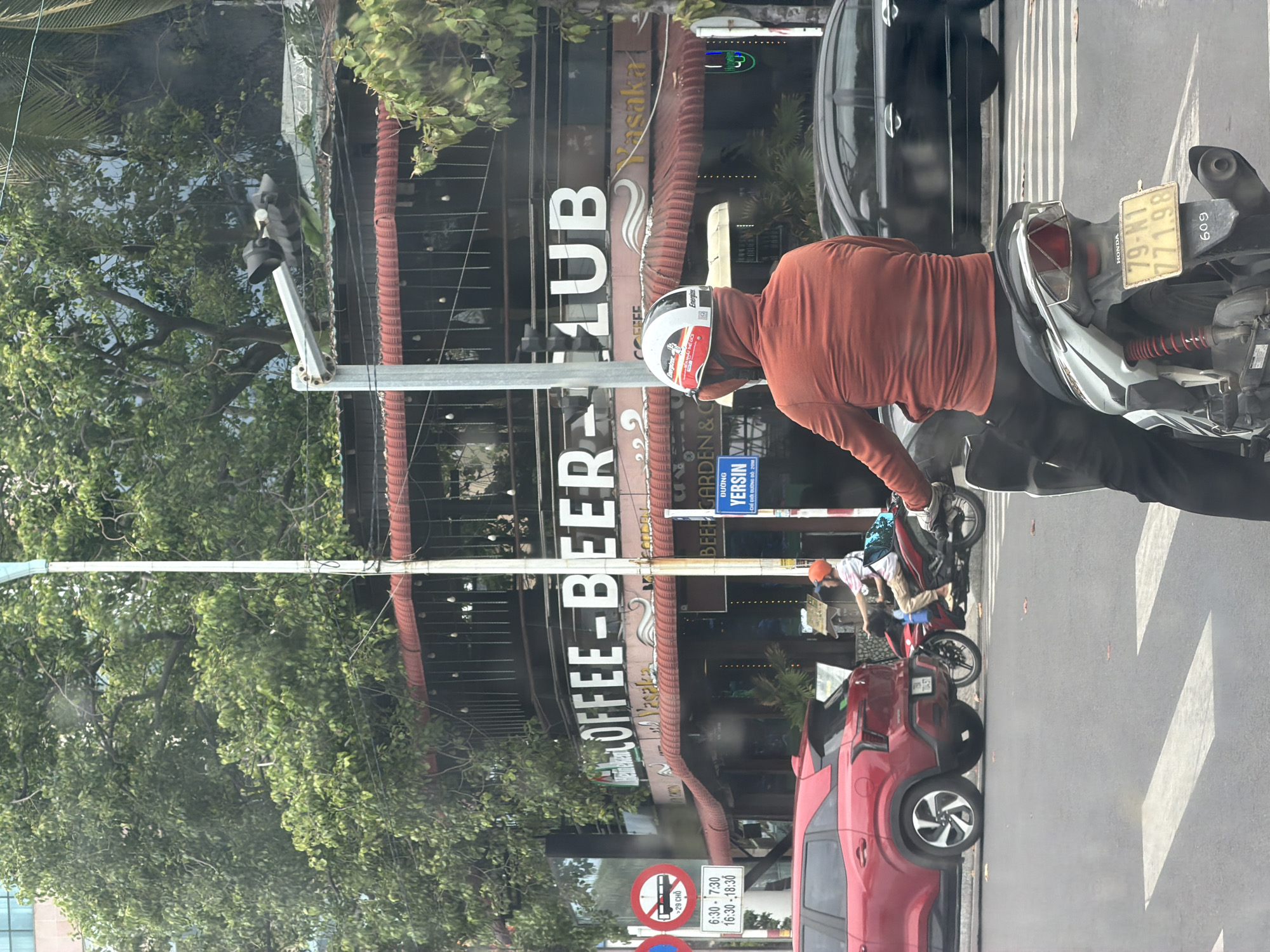 A busy street scene is captured in a vertical, eye-level shot. A man wearing a red long-sleeved shirt and a white helmet with an Energizer logo is on a motorbike, with the license plate 79-N1 771.98 visible. The motorbike's make is Honda, with a number "609" displayed. Behind him, a red car is parked. Further down the street, there is another motorbike with two individuals on it.
The backdrop features a building with the words "COFFEE-BEER CLUB" and "Yasaka" prominently displayed. A sign points towards Yersin Street and includes regulations for traffic restrictions. Lush green trees are present on the left side of the image. The scene appears to be in Nha Trang Ward, Vietnam.