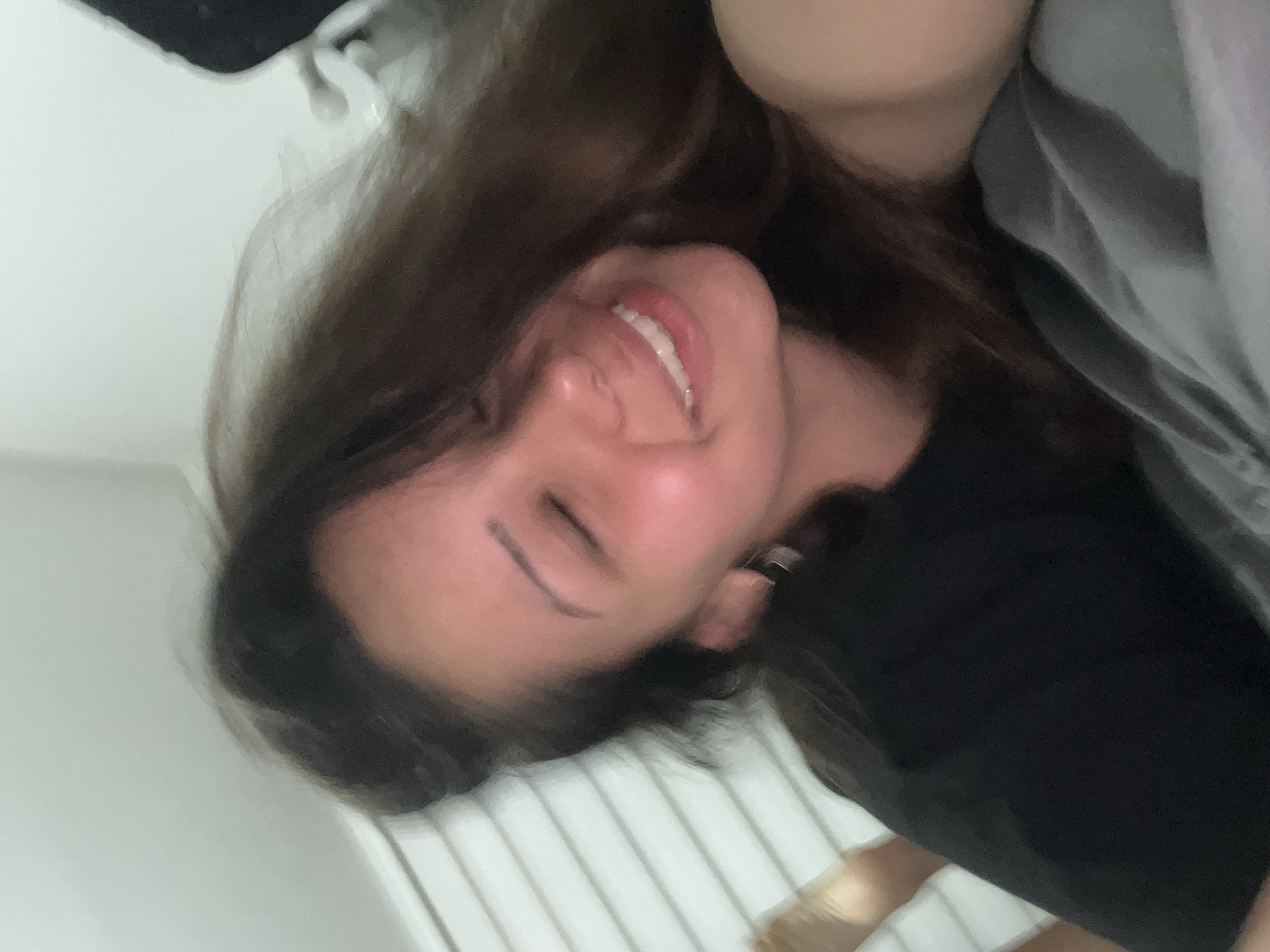 A woman with long, dark brown hair smiles with her eyes closed while lying on a gray blanket. She is wearing a black t-shirt. The background shows white horizontal blinds. The image was taken in Zlatograd, Bulgaria.