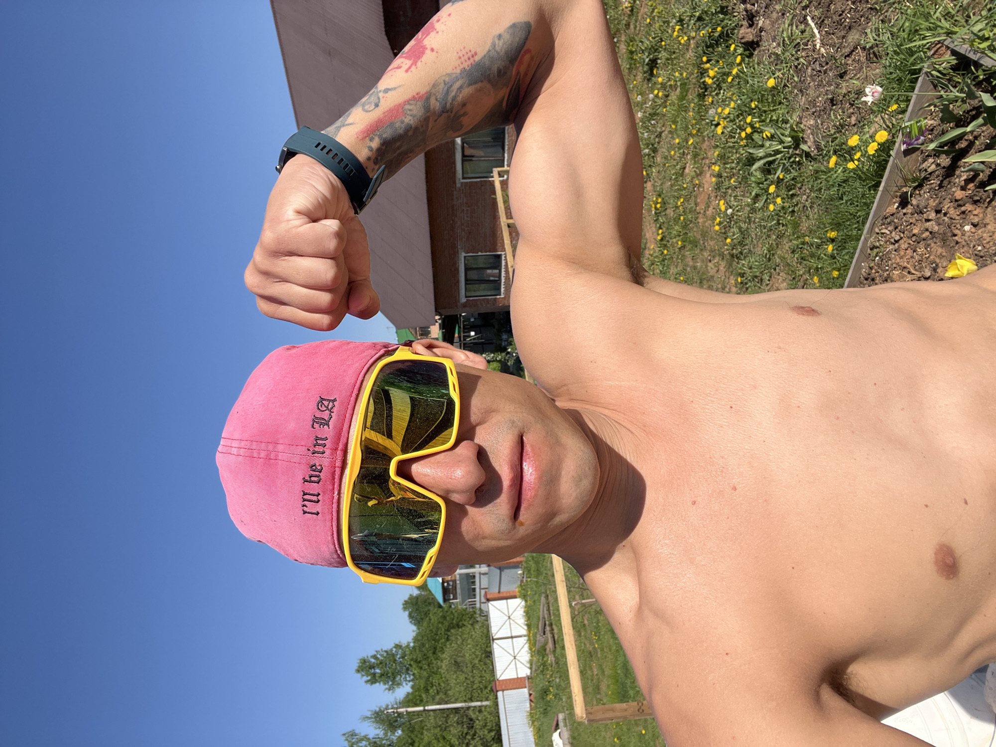 A shirtless man with a pink baseball cap and yellow sunglasses is posing outside on a sunny day. The hat has the phrase "I'll be in LA" written on it. He is flexing his left bicep, which has a tattoo on the upper arm. He is wearing a green smart watch on his left wrist. The location appears to be a yard with grass and dandelions, possibly in the city of Chaikovsky, Russia, based on contextual information. A building is visible in the background, and a small garden bed or wooden structure is present in the foreground.