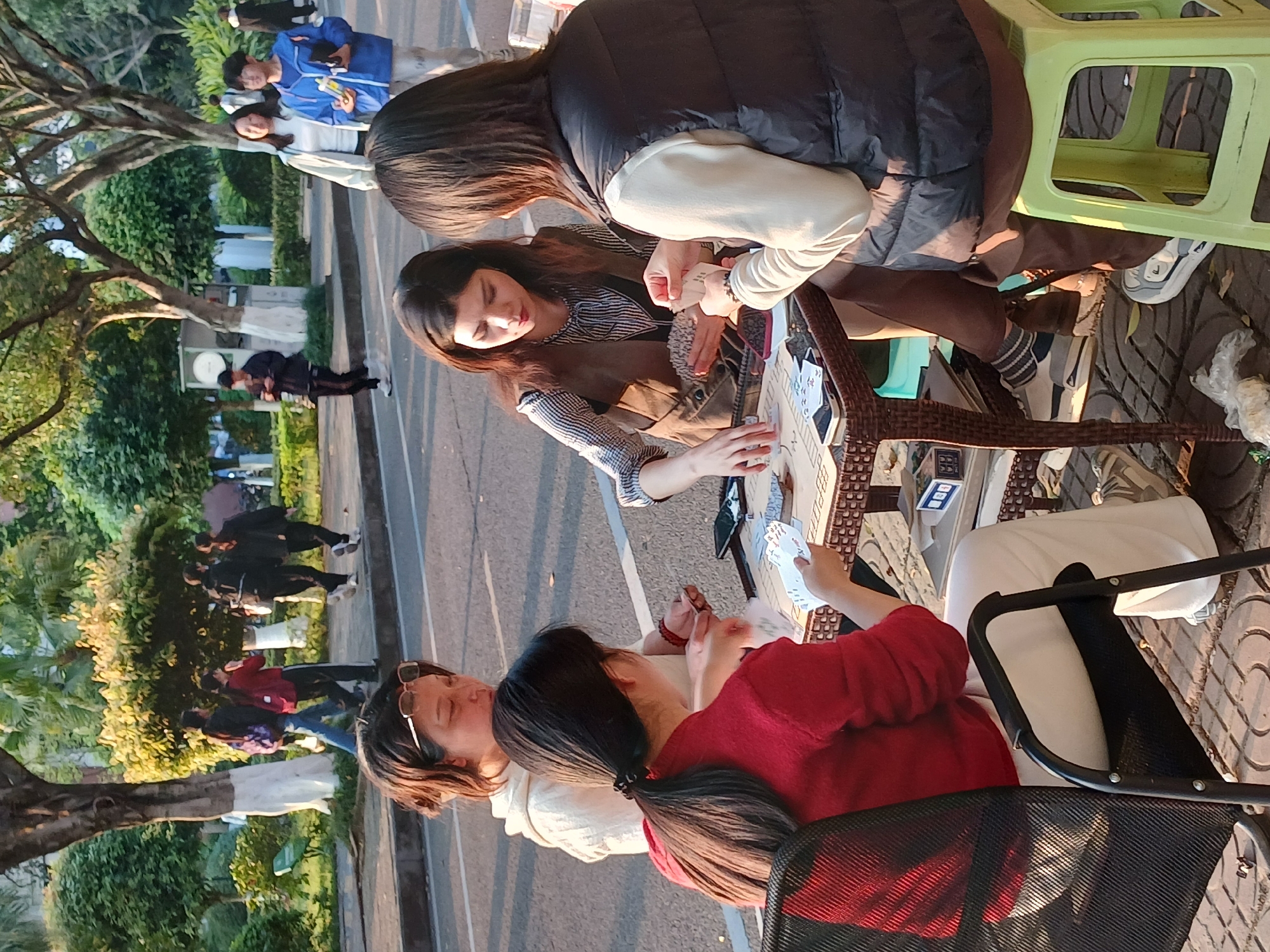 An outdoor scene in the Chongqing High-tech Zone, China, depicts four adult females engaged in a card game on a paved sidewalk. Direct sunlight illuminates the scene, casting shadows.
In the immediate foreground, a small, square, dark brown wicker-patterned table serves as the focal point. Around it are seated four women:
1. A woman with dark hair tied in a ponytail, wearing a red long-sleeved top and white pants, is positioned with her back to the camera. She sits on a black office-style chair and holds cards in her left hand.
2. To her right, a woman with dark hair and sunglasses pushed onto her head, wearing a light-colored long-sleeved garment, is partially visible, looking down towards the table.
3. Directly opposite the first woman, a third woman with dark hair, wearing a striped long-sleeved shirt and a brown sleeveless vest, actively manipulates cards on the table and in her left hand.
4. To her right, a fourth woman with dark hair, wearing a cream-colored long-sleeved top and a black padded vest, sits on a bright lime-green plastic stool, holding cards in both hands.
Multiple playing cards are scattered across the table surface, alongside a dark-colored mobile phone. The ground beneath the table consists of patterned paving stones, with a discarded clear plastic bag visible.
Behind the card game setup, an asphalt road separates the foreground activity from a tree-lined sidewalk. Several pedestrians are present on the far sidewalk: a group of four, including one individual in a red jacket and another with a backpack, walks towards the left, while a group of three, including one in a light jacket and another in a blue jacket, walks towards the right. The sidewalk is bordered by trees featuring trunks painted white at their bases and dense green foliage. A white utility box is discernible further along the sidewalk.