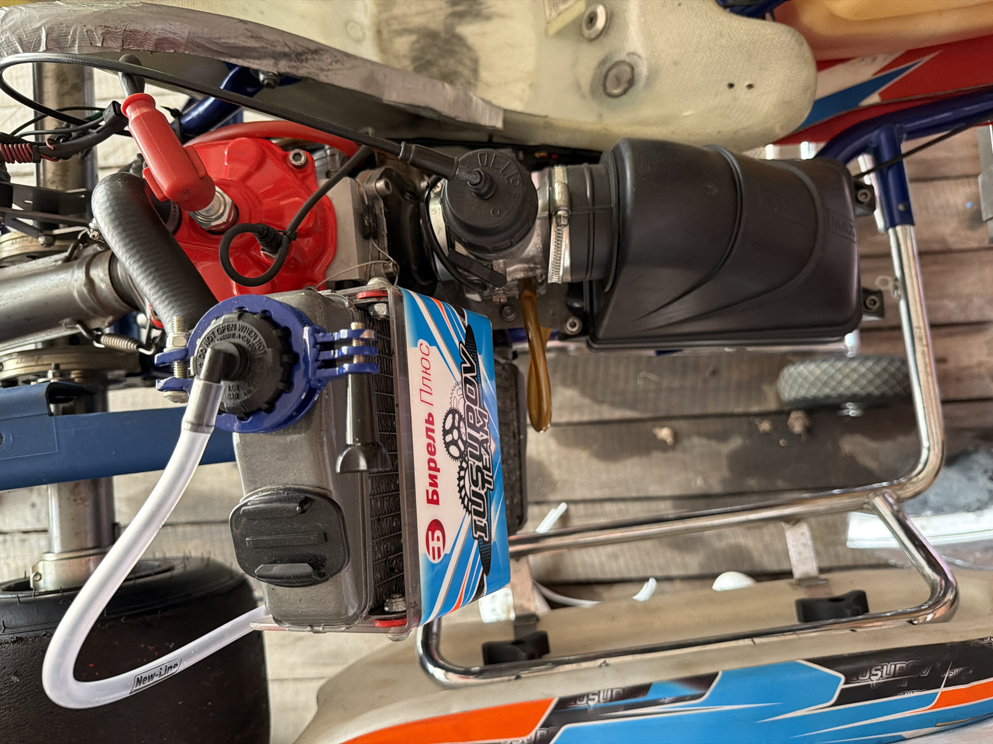 This is a close-up, overhead shot of a racing go-kart engine assembly, likely taken in an outdoor or garage setting, possibly in Sukhaya, Russia. The focus is on the engine components, including a fuel tank with branding that reads "БИЯРЕЛЬ ПЛЮС" and "IUSUPOL TEAM". A clear plastic hose is visible, curving from the top left corner towards the engine, with "New-Line" printed on it. The engine features red and blue parts, a black ribbed hose, and a red spark plug cap. A black rectangular reservoir is mounted below the engine. The go-kart bodywork, featuring blue and white graphics with orange accents, is partially visible on the left side. The background consists of wooden planks, suggesting a workshop or track environment. There are no people or animals visible in the image. The lighting appears to be natural, possibly from an overcast sky or ambient light in a workshop.