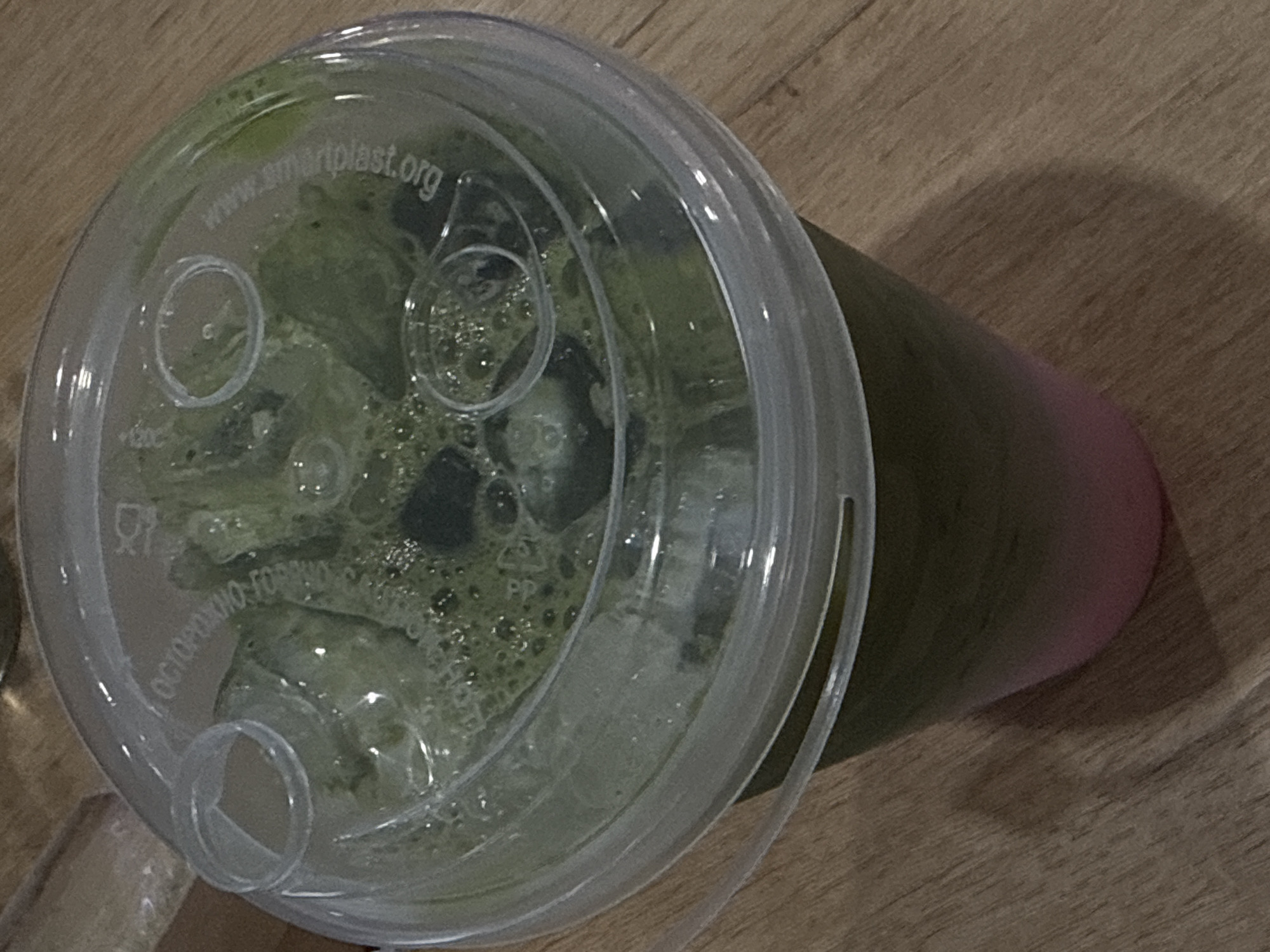 The image shows a close-up of a clear plastic cup filled with a green beverage and ice. The cup has a clear plastic lid with a straw hole. The beverage appears to be a matcha drink with a layer of pink liquid at the bottom. The cup is placed on a wooden surface, likely a table. There is some text embossed on the lid, which is difficult to read clearly due to the condensation and lighting. The background is out of focus, showing more of the wooden surface. The image does not provide any information about the location in Lobacheve, Ukraine, or any people, actions, or interactions.