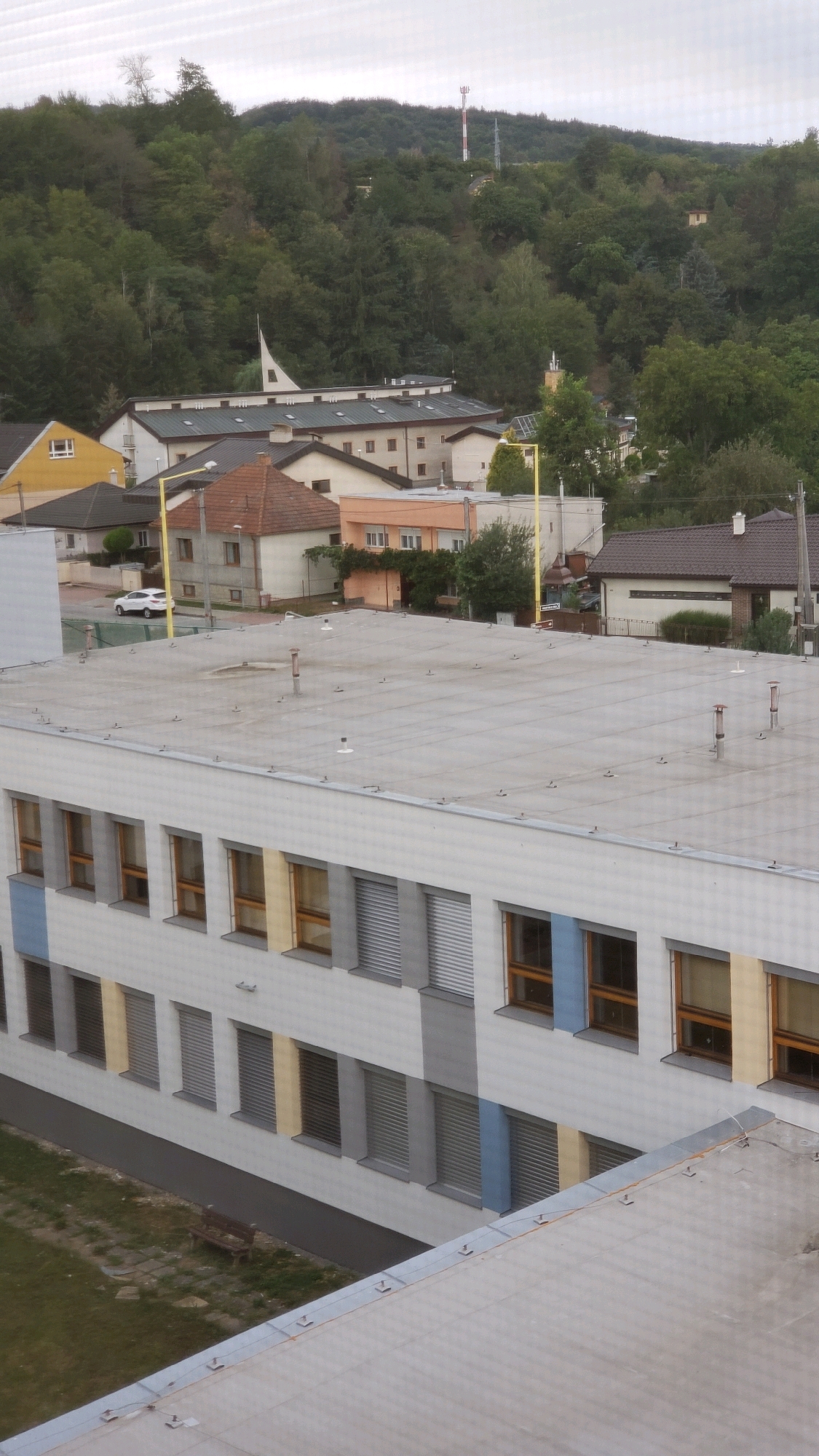 The image shows a view of the town of Prešov, Slovakia, from a high vantage point. In the foreground, there is the flat, gray roof of a modern building, likely a school or an office building, with several vent pipes and small antennas. The building's facade is visible, featuring rows of windows with a mix of wooden frames and metal blinds, and sections of the wall are painted in white, gray, yellow, and blue. Below the building, a patch of grass with a lone wooden bench can be seen.
In the mid-ground, several smaller buildings with pitched roofs are clustered together, some with reddish-brown tiles and others with gray roofing. One prominent structure has a distinctive white, modern, curved roof, possibly a church or a cultural building. Further back, more residential houses and buildings are scattered amongst lush green trees.
In the background, a densely forested hill rises, with a tall red and white telecommunications tower visible near its summit. The sky is overcast, casting a muted light over the entire scene. The presence of a white car on a road in the mid-ground suggests ongoing activity within the town.