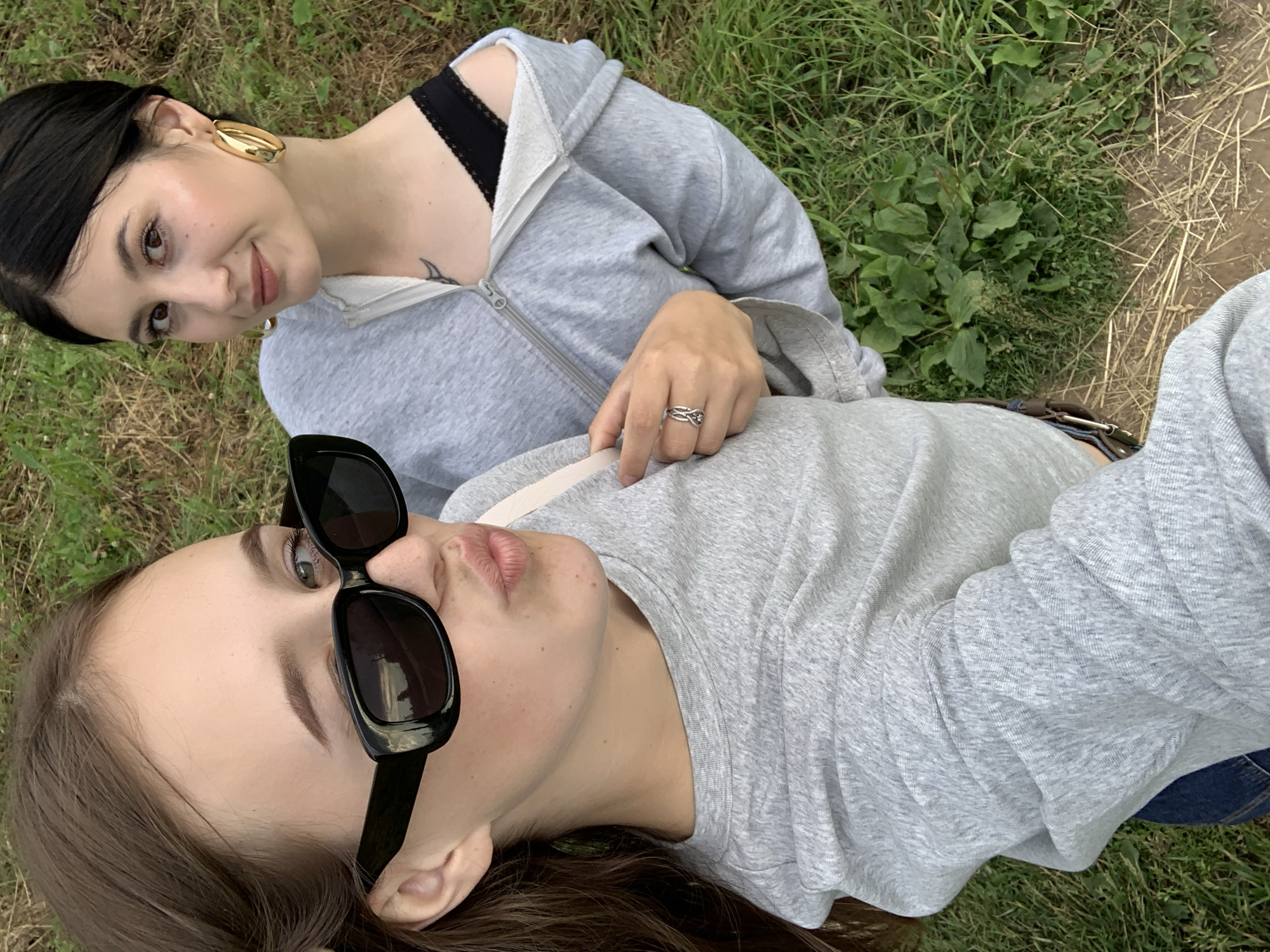 Two young women are captured in a high-angle selfie outdoors. The woman in the foreground, on the left, is wearing a grey long-sleeved shirt and black sunglasses, with her lips pursed. The woman on the right is wearing a grey zip-up hoodie, partially unzipped to reveal a black lace bra. She has dark hair pulled back and is wearing large gold hoop earrings. Both women appear to be in a grassy area with some dry grass and dirt visible. The location is described as the city of Fokino, Russia.