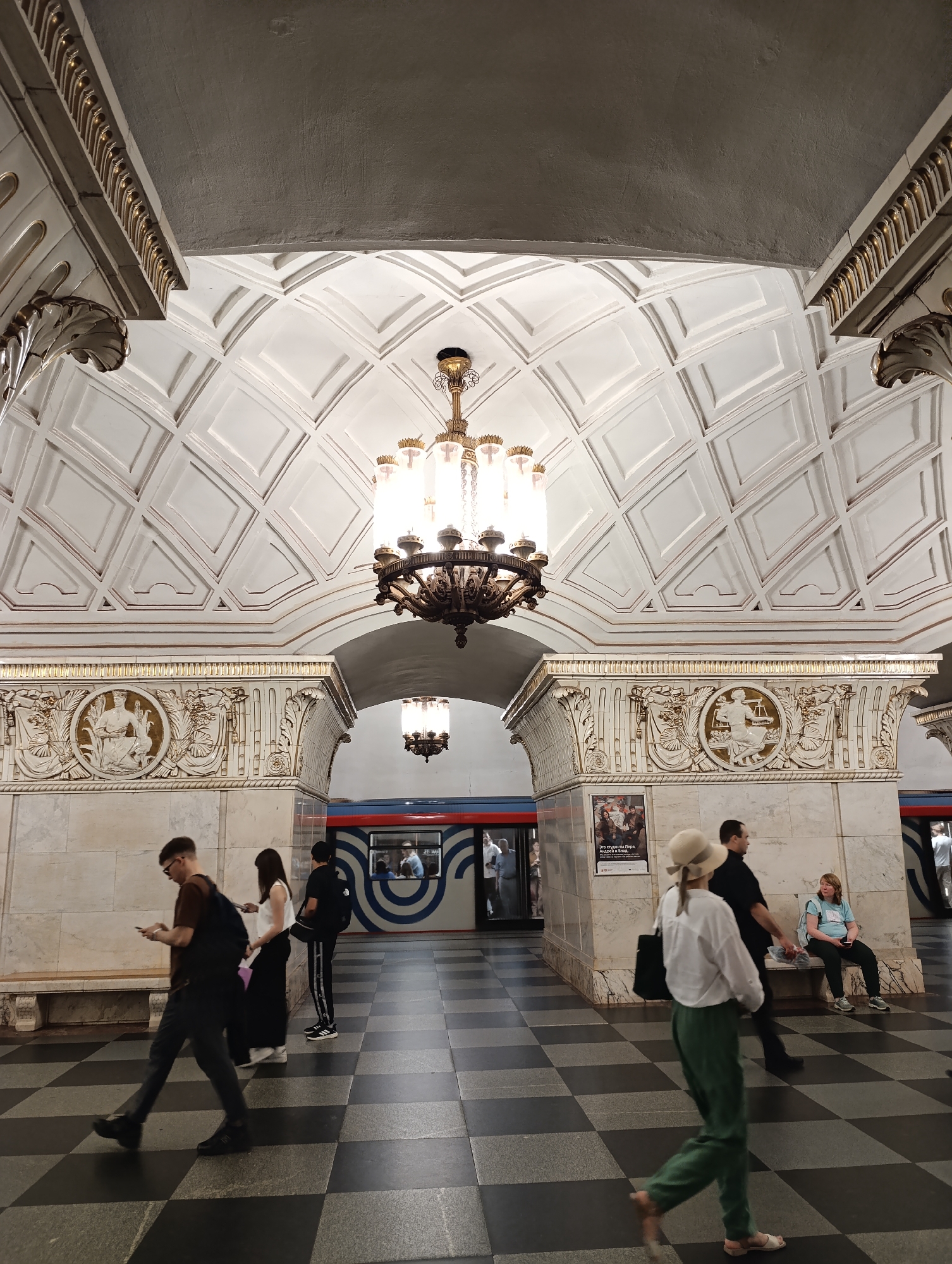 The image displays a grand and ornate interior of a subway station, characteristic of the Moscow Metro. The high ceiling is a striking feature, with a white, geometrically coffered pattern accentuated by subtle gilded lines. Large, elaborate bronze chandeliers with multiple lights hang prominently from the center, illuminating the space with a warm glow.
The station walls are clad in light-colored, possibly marble, panels and topped with richly decorated friezes. These friezes feature intricate relief sculptures, including figures, one of which appears to be holding scales, set within circular medallions and flanked by stylized foliage and vertical patterns, all highlighted with gold accents. Large, square columns or support structures are also adorned with these decorative elements.
The floor contrasts sharply with the light upper structures, laid out in a distinct black and white checkerboard pattern. Several people are present on the platform: in the foreground, a man in a dark shirt and backpack walks left while looking at his phone, followed by a woman in a white top. On the right, a woman in a straw hat and green pants walks towards the viewer, and further back, another woman sits on a bench. Through an archway in the background, a modern Moscow Metro train with its distinctive blue, white, and red stripes is visible at the platform, its doors open, suggesting it is either arriving or departing.
On a column to the right, a poster with Cyrillic text is visible, reading: "Это студенты Петя, Андрей и Дима." (These are students Petya, Andrey and Dima.) The overall atmosphere is that of a bustling yet architecturally impressive public transport hub. The lighting is entirely artificial, consistent with an underground setting.