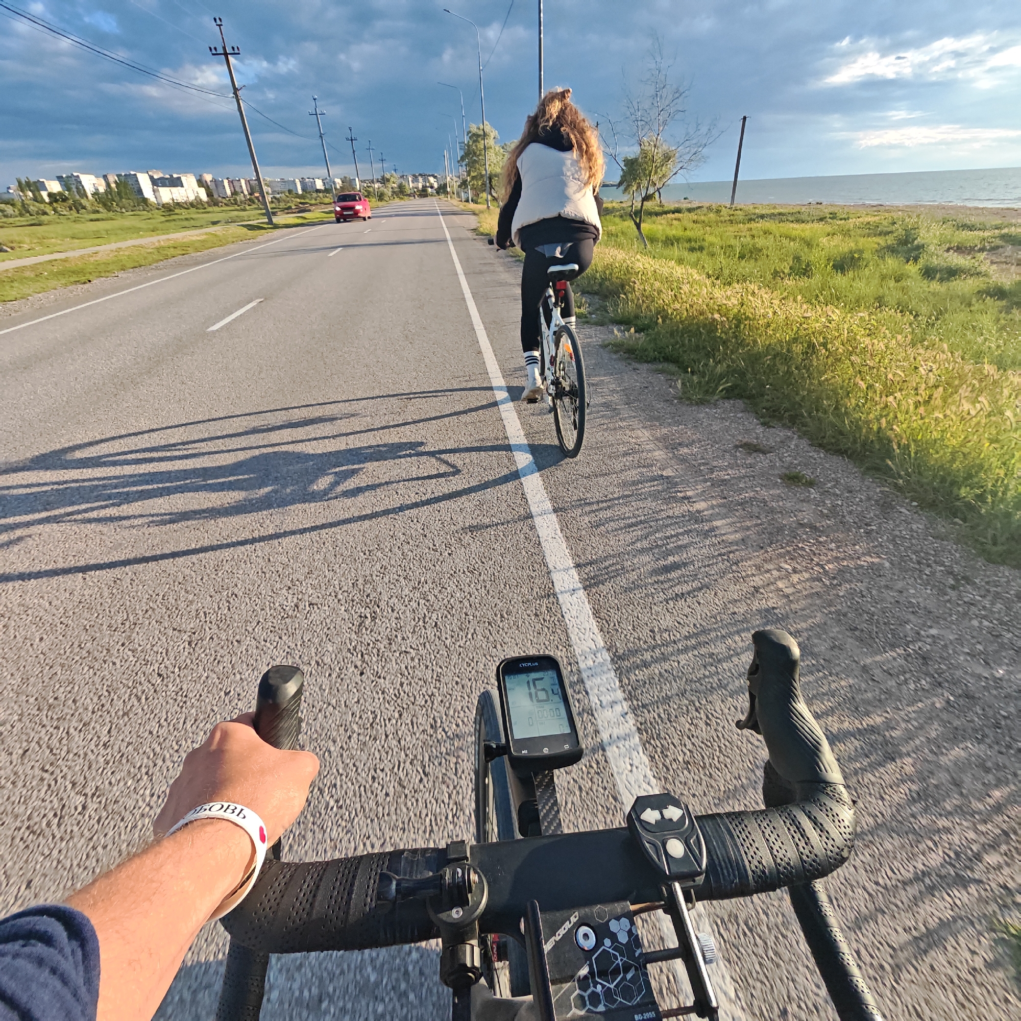 The image captures a first-person view from a person riding a bicycle, following another cyclist on a paved road. In the foreground, the photographer's right arm and hand are visible, gripping black handlebars. A white wristband with partial text "БОВЬ" (likely "ЛЮБОВЬ" meaning "love") is on the wrist. A black bicycle computer, labeled "CYCPLUS," is mounted on the handlebars, displaying "16.4" (presumably speed) and "00:00." Additional bike accessories are visible, including one marked "PENGGUO RQ-2055."
Ahead, another person on a bicycle is riding away, slightly to the right of the road's white dividing line. This cyclist has long, curly, light brown hair and is wearing a black top, a white puffer vest, black leggings, and white socks with black stripes. Long shadows of the cyclists stretch across the road, indicating either early morning or late afternoon light.
To the left of the road, power lines and utility poles line the route, with distant residential buildings visible on the horizon. A red car is seen approaching in the opposite lane. To the right of the road, a strip of green grass and foliage separates the road from a large body of water, likely the Azov Sea, under a partly cloudy sky. The setting suggests an outdoor recreational activity in Мысовское сельское поселение, Ukraine.