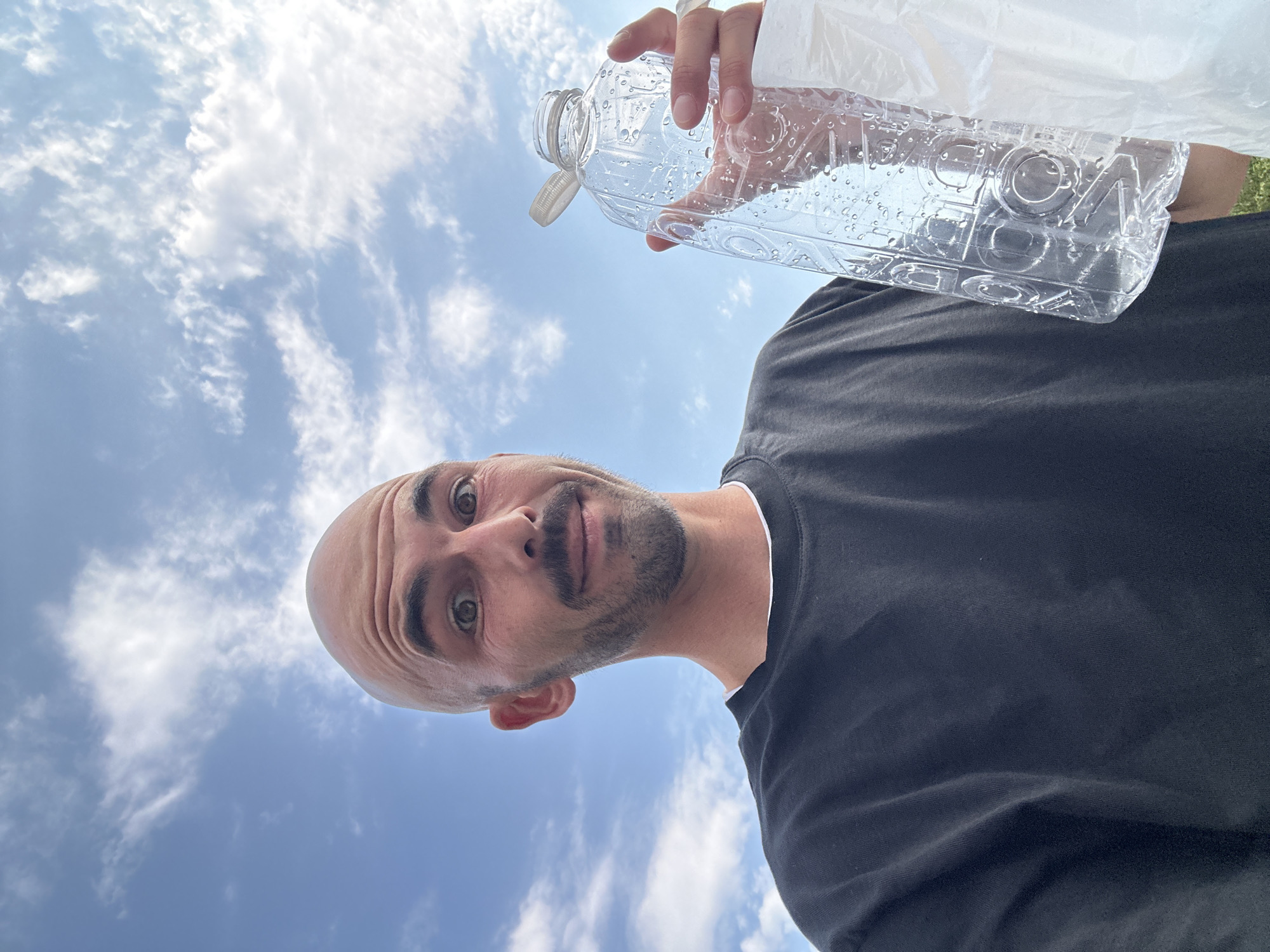 An individual is photographed from a low angle, holding an empty plastic water bottle filled with water droplets. The person, who is bald with a goatee and mustache, wears a black t-shirt with a white collar visible. The background is a bright blue sky with scattered white clouds. The image is shot outdoors, and the location is identified as Drobnići, Montenegro. The person is looking towards the camera with a surprised expression. The bottle has raised lettering, though the text is partially obscured by the water droplets. The individual's left hand is visible, holding the bottle, and a white plastic bag is held in their right hand.