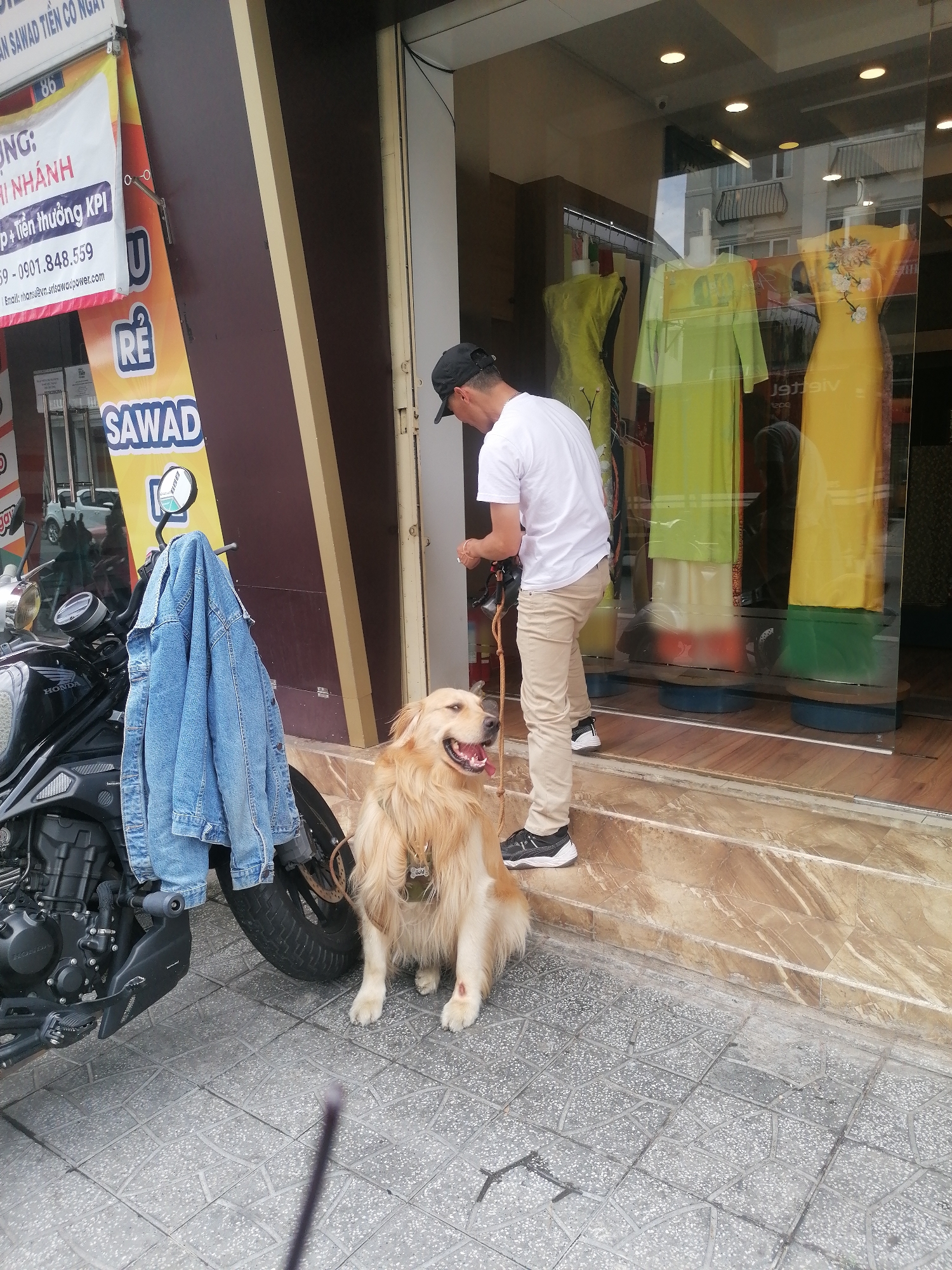 A full outdoor shot in Vietnam, in the city of Da Lat shows a man and dog outside of a shop. The man is fair-skinned and wears a black baseball cap, a white t-shirt, and tan slacks and black sneakers. He is turned towards the glass shopfront. A large golden retriever dog sits in front of him. The dog has a light-colored coat and has a bloody patch on its front leg. Attached to the front tire of a black motorcycle is a jean jacket. The storefront is glass and marble. Three yellow dresses hang in the window. The motorcycle is parked in front of the storefront. A sign is visible above the motorcycle.