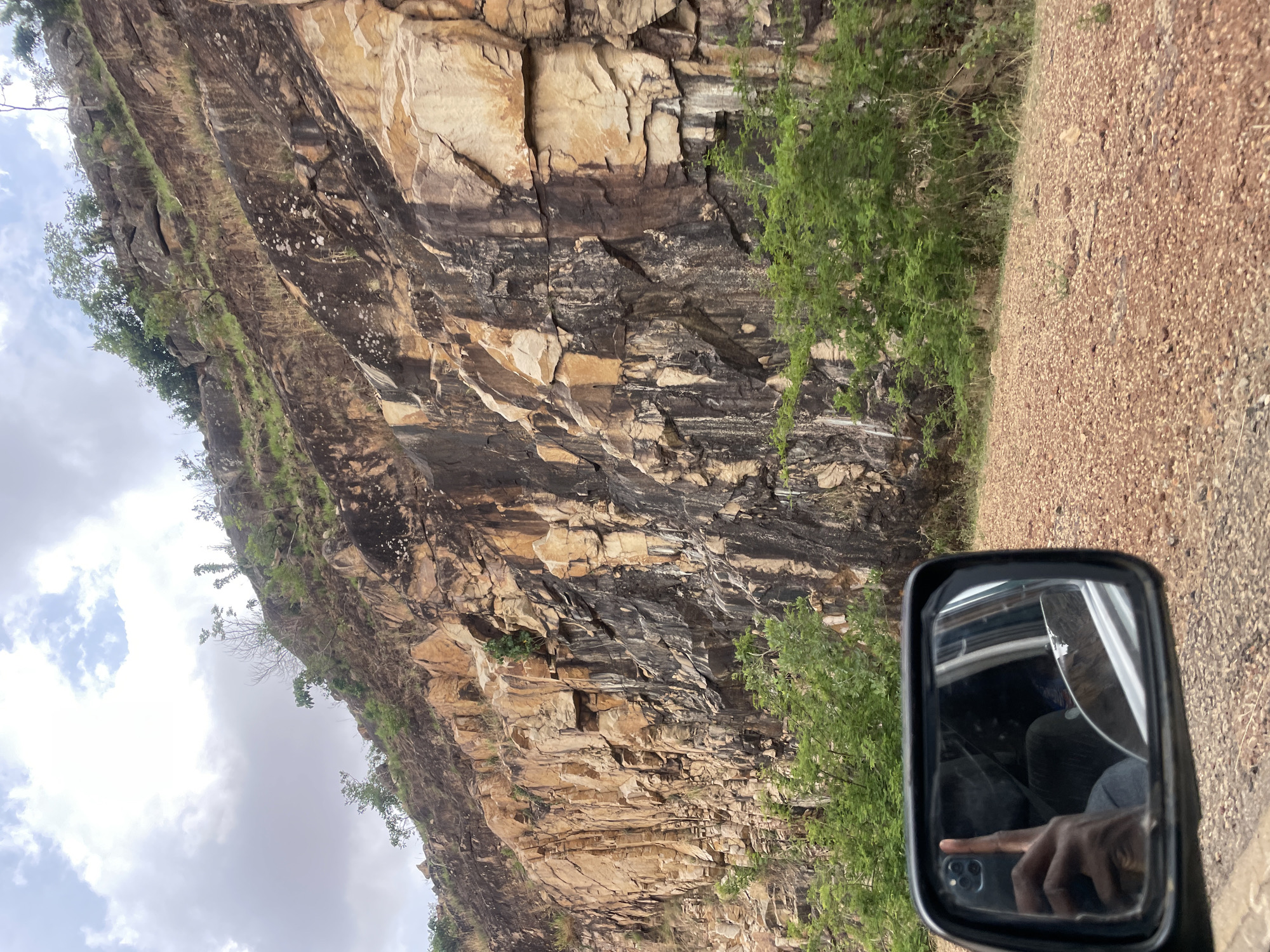 The image depicts a vertical rock cliff with alternating bands of dark and light colored rock, partially covered with greenery at its base and some vegetation along its top edge. The cliff appears to be located alongside a dirt road, as suggested by the surface texture of the foreground on the right side of the image. The view is likely taken from a vehicle. Part of a side mirror is visible in the lower right corner, showing a person inside holding a phone taking a photo. This appears to be a location in Vom, Nigeria.