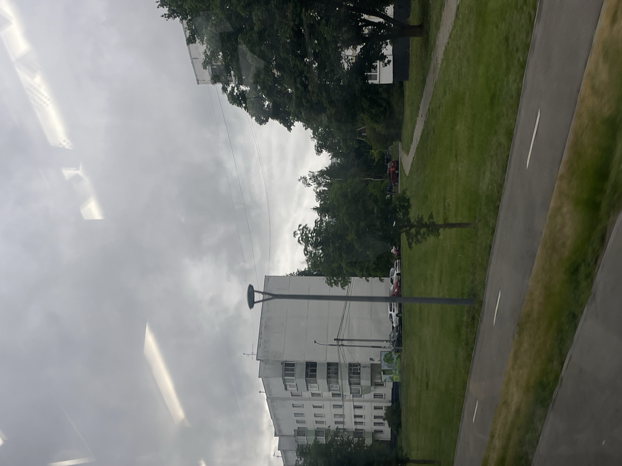 The image depicts a grey, overcast sky seen through a window, with reflections of ceiling lights. Outside, a residential building with multiple stories and balconies is visible, surrounded by trees. A paved path and a grassy area are also present. Several cars are parked along the side of the road near the building. The overall scene suggests a cloudy day in a residential area, likely in Yakovlevskoye, Russia, as indicated by the location context. The image is taken from an interior perspective looking outwards, and the orientation of the image is vertical. No people are visible, and there is no discernible text.