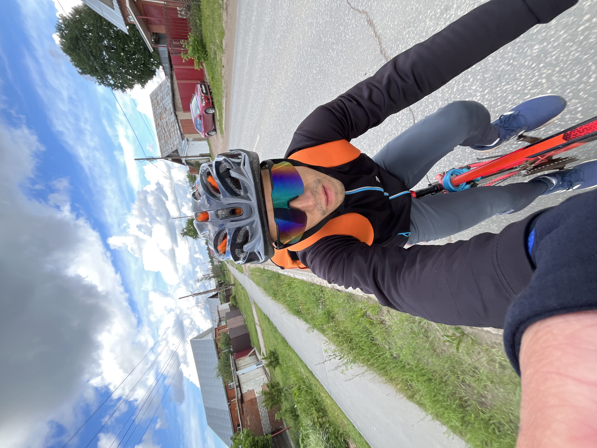 The image captures a dynamic, high-angle selfie taken by a male cyclist during daytime in Chaikovsky, Russia. The man is positioned in the foreground, wearing a grey cycling helmet with orange details, and reflective, multi-colored sunglasses that obscure his eyes. He is dressed in a black long-sleeved cycling top with prominent orange panels on the shoulders and chest, grey trousers, and blue athletic shoes.
He is actively riding a bicycle, part of which is visible with a red frame and a blue cable or lock wrapped around the front. The perspective is from the rider's viewpoint, looking slightly down and forward.
The scene unfolds along a paved road, which occupies the central and left portions of the background. A red car is parked on the far side of the road. To the right, a concrete sidewalk runs parallel to the road, bordered by vibrant green grass. Beyond the road and sidewalk, various houses, characterized by different architectural styles including brick and simple siding, line the street. Overhead utility wires traverse the sky, suggesting an established residential or semi-urban environment.
The sky is a notable element, featuring a mix of bright white cumulus clouds against patches of clear blue, indicating fair weather conditions. The overall composition is tilted, imparting a sense of motion and the immersive experience of cycling. The man's expression is neutral as he captures the moment.