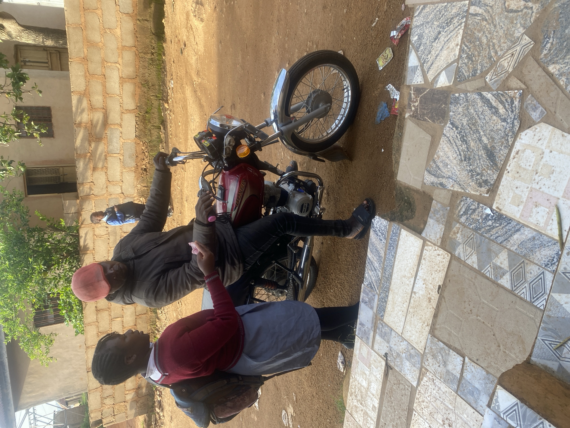 In Gura Top, Nigeria, a young Black female student wearing a red sweater, grey skirt, and black stockings stands beside a red motorcycle. A Black male, wearing a dark jacket and a pink cap, sits on the motorcycle, handing the student a pink item, possibly money or candy. The student has a backpack on her left shoulder. In the background, a brick wall separates the scene from a building with a light-colored facade and two doors. Another young Black male in a blue jacket is visible further in the background. The ground is dirt with scattered debris, and the foreground features a tiled pathway.