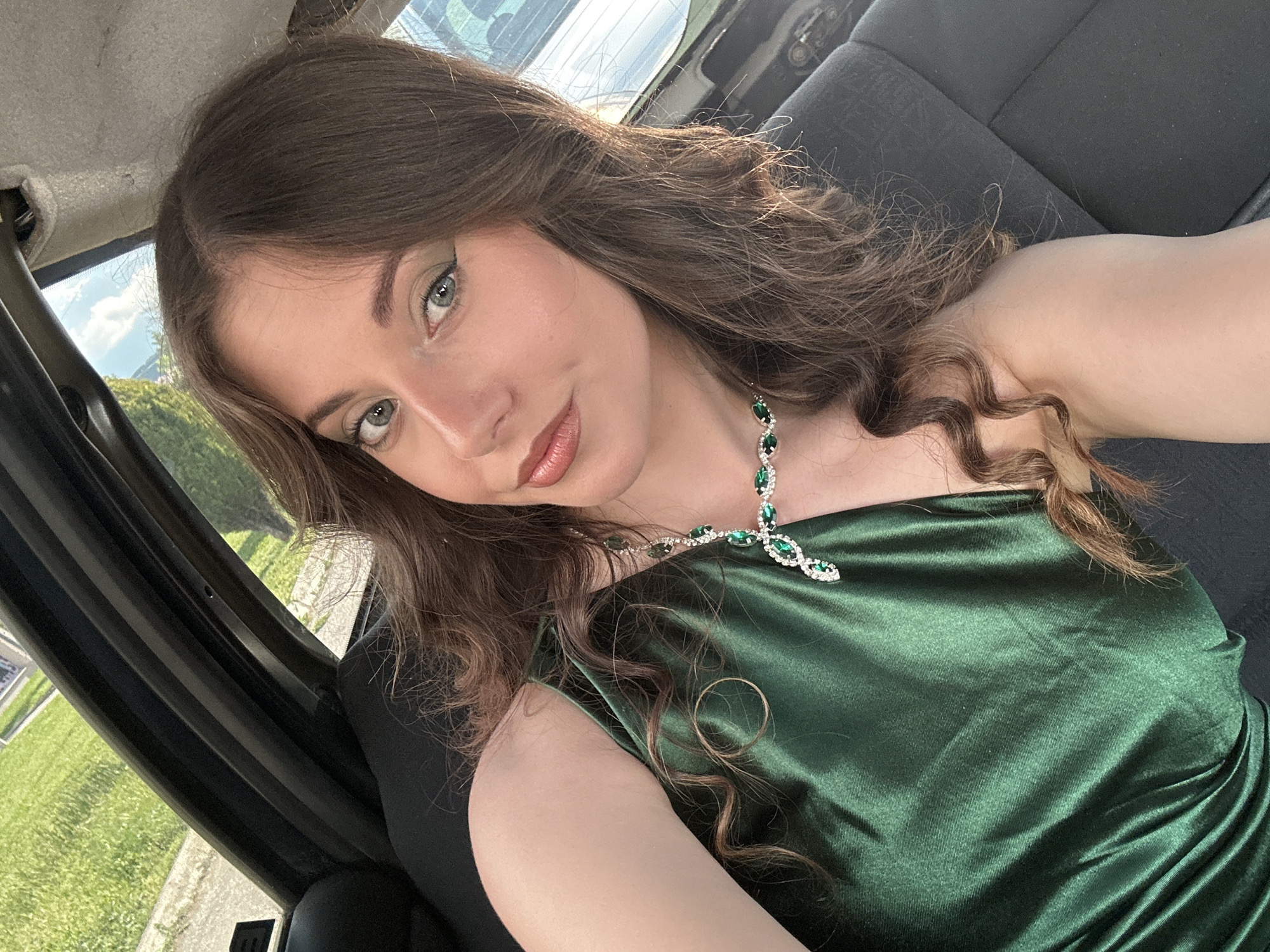 A fair-skinned woman with brown, wavy hair is shown in a medium close-up selfie taken inside a car. She is wearing a green, shiny dress with a green gemstone necklace. The car's interior is visible, including the black seats and the car door frame. Through the car window, a glimpse of a green landscape and a building can be seen. The location is unknown.