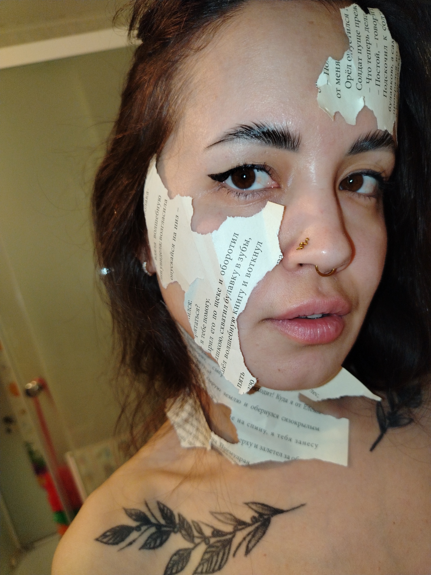 A woman with dark hair and a leaf tattoo on her upper chest poses indoors. Torn pieces of a page with Russian text cover parts of her face. The text includes phrases like "от меня" (from me), "волшебную книгу" (magic book), and "я тебе помогу" (I will help you). The woman has a nose ring and defined eyeliner. A red toothbrush and other bathroom items are visible in the background. The location of the image is not specified, Noviy is not in Russia.