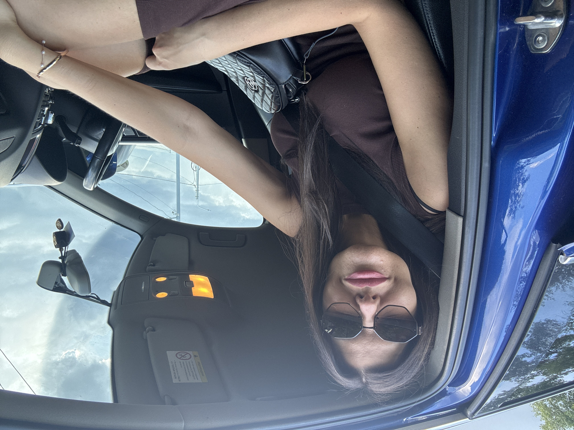 In Chaikovsky, Russia, a woman with dark hair and sunglasses is pictured inside a blue car, wearing a brown sleeveless dress and a black purse slung across her body. The car's interior is visible, including the steering wheel, dashboard, and interior lights. The sky outside is cloudy. The woman is leaning against the car door.