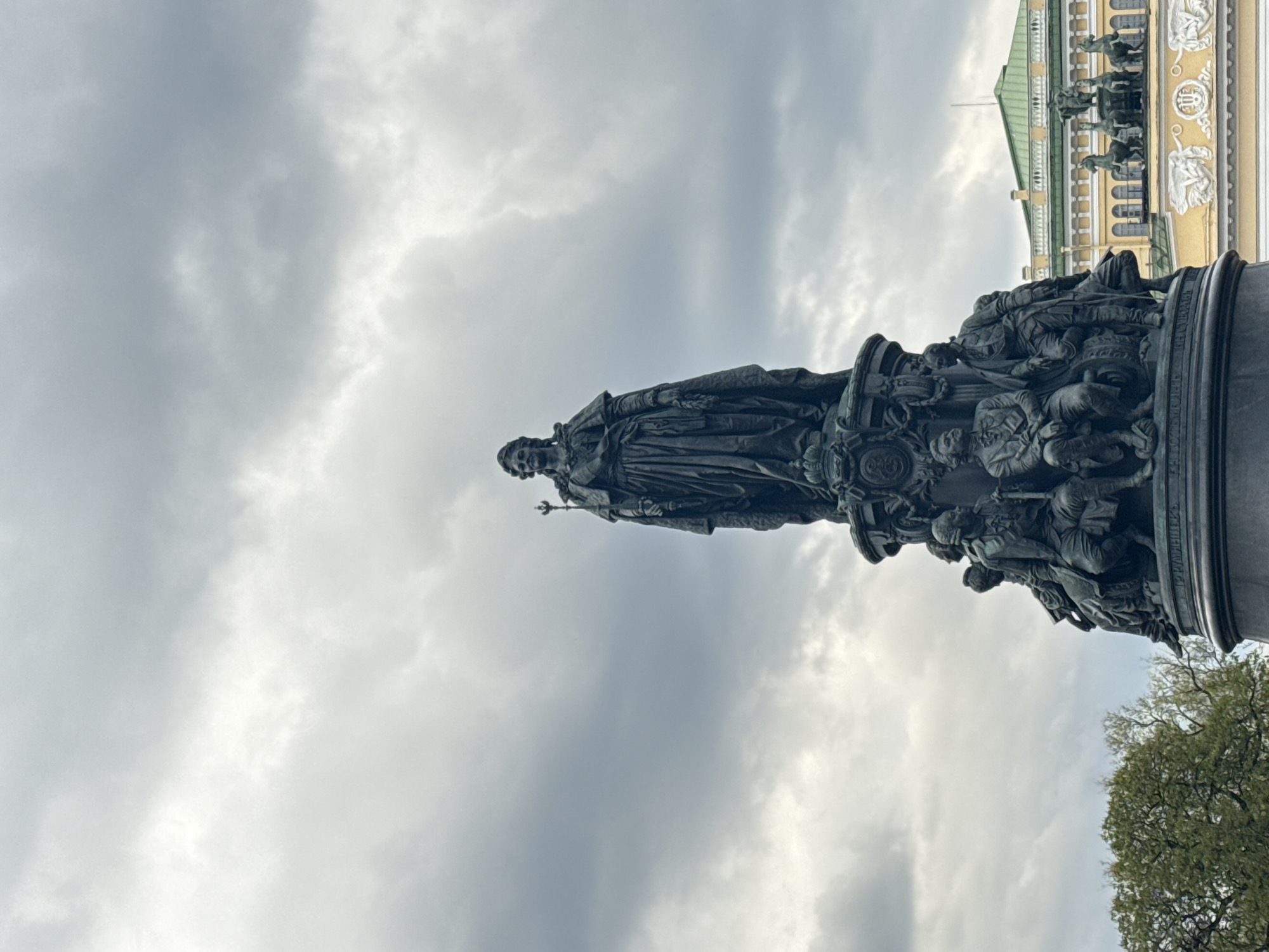 The image depicts the Catherine the Great Monument in Saint Petersburg, Russia. The bronze statue of Catherine is centrally positioned, standing tall against a backdrop of cloudy skies. The monument is surrounded by other historical figures at its base. Adjacent to the monument, a building with classical architecture, including columns and a green roof, is visible. Some greenery is also seen to the side, indicating the presence of trees in the vicinity.