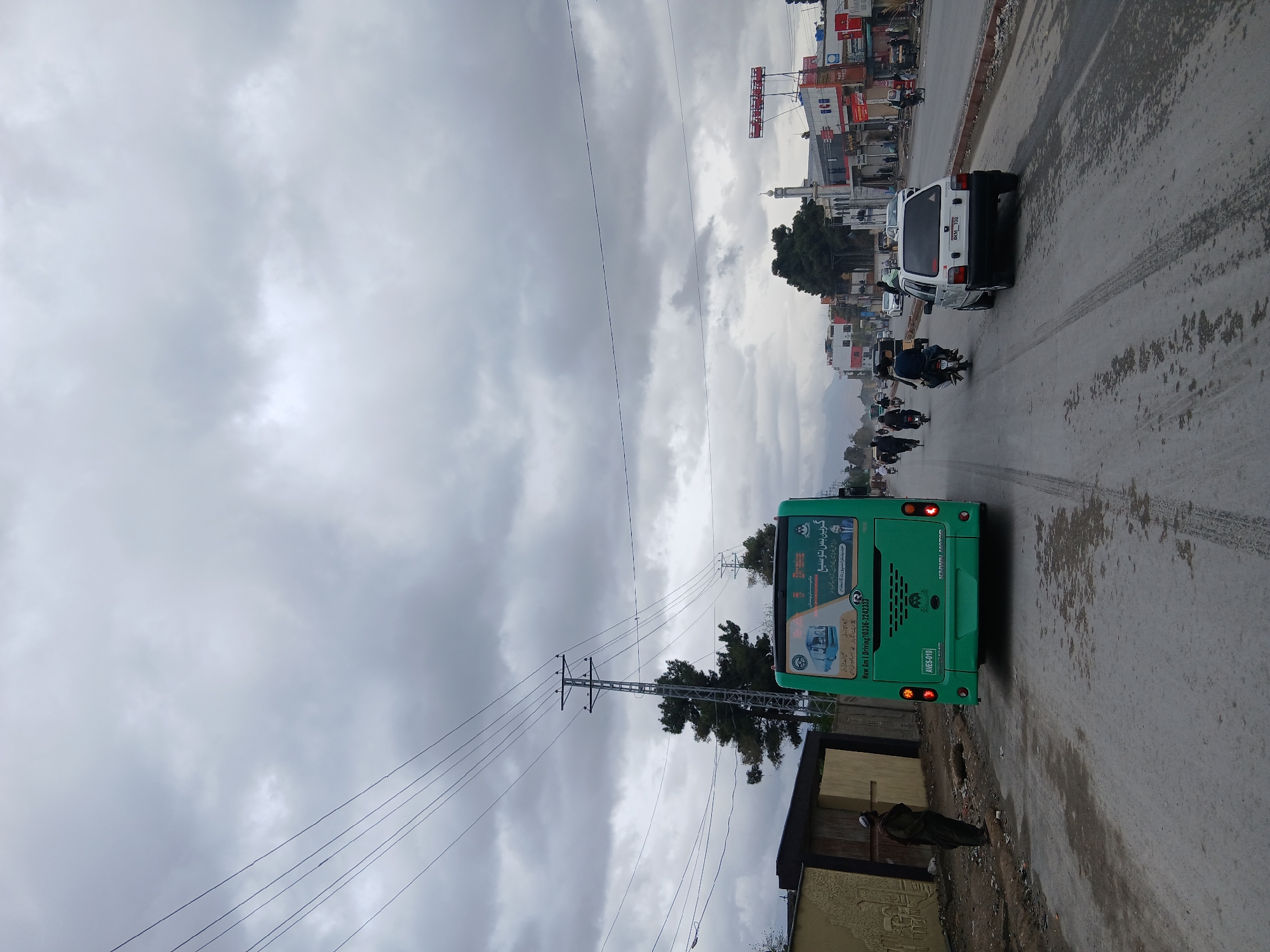 The image shows a street scene in Quetta City Tehsil, Pakistan, under a cloudy sky. A green bus occupies the right side of the frame, with its rear facing the camera. The bus has advertisements and signage on its back. In front of the bus, a white van and several motorcycles are visible on the road. Along the roadside, a building and a utility pole are present. A person is standing near the building. In the background, there are buildings and a minaret, indicating an urban environment. The road appears wet, suggesting recent or ongoing precipitation.