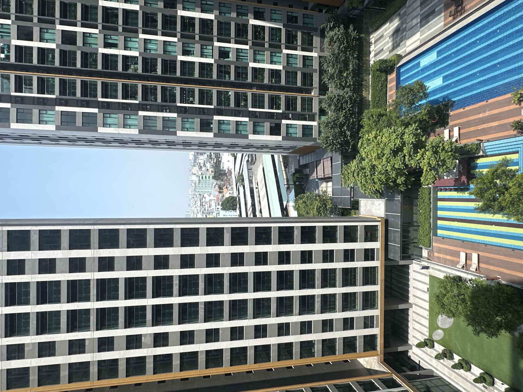 An aerial view from a high vantage point depicts a modern urban residential complex in Thủ Đức, Vietnam, situated between two multi-story buildings. The central ground level features an unoccupied recreational area. On the right, a large, rectangular swimming pool displays blue tiling with lighter blue lane lines. Adjacent wooden-patterned decking includes two light-colored loungers. To the left of the pool, a distinct area with blue, yellow, and green vertical stripes incorporates integrated steps, suggesting a decorative water feature or paved zone. Green spaces, including sections of artificial turf with three white circular ground markings, and mature trees, are interspersed with geometrically patterned grey paved walkways. No individuals are visible in the recreational areas or on the building facades. The two flanking high-rise buildings present modern architectural styles: the left building features a grey grid pattern with green-tinted balcony railings and a vertical yellow-brown stripe; the right building displays a darker grey base with white vertical and horizontal accents, similar green-tinted windows/balconies, and a central vertical yellow-brown stripe. The background reveals an urban landscape of lower-rise buildings, including a large light-green roofed structure, extending to a distant city skyline under a clear blue sky.
