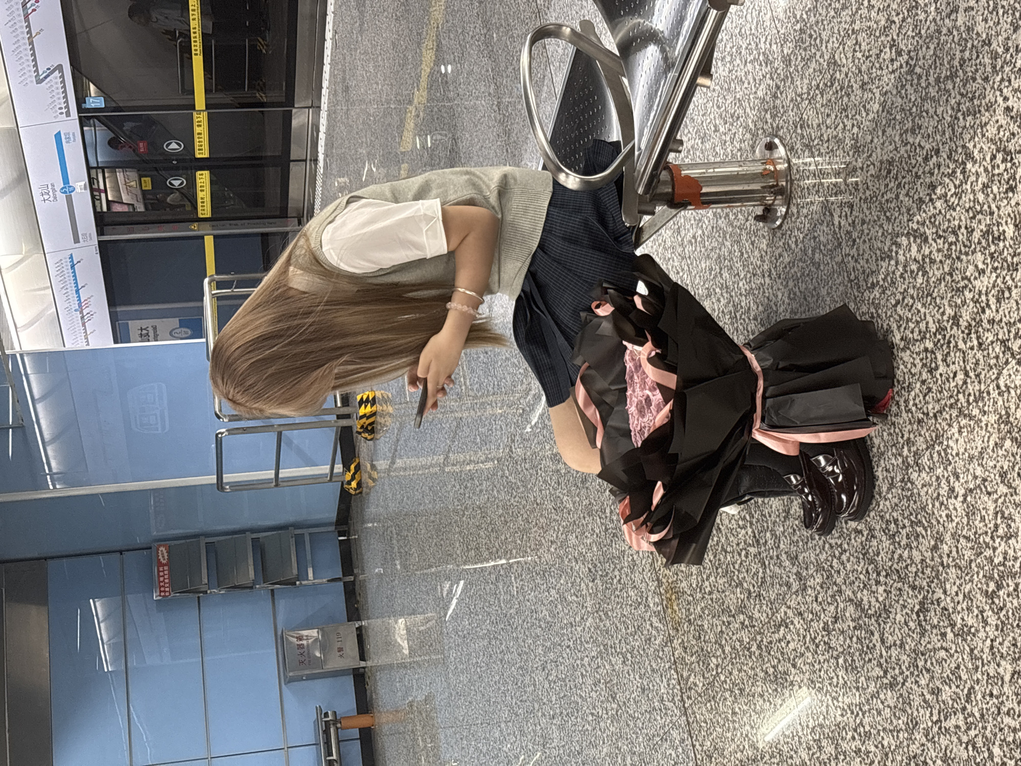 The image shows a person standing at a subway station platform in Liangjiang, China. The person is fair-skinned, with long blonde hair, and is wearing a white shirt, a grey sweater, and a blue pleated skirt. They are holding a cell phone in their hands, looking down at it. Leaning against a bench are a black bag and a bouquet of black and pink flowers. The subway platform is made of grey tiled floor, and the walls behind the platform are covered in light-blue tiles. There is a subway car behind a transparent security panel.