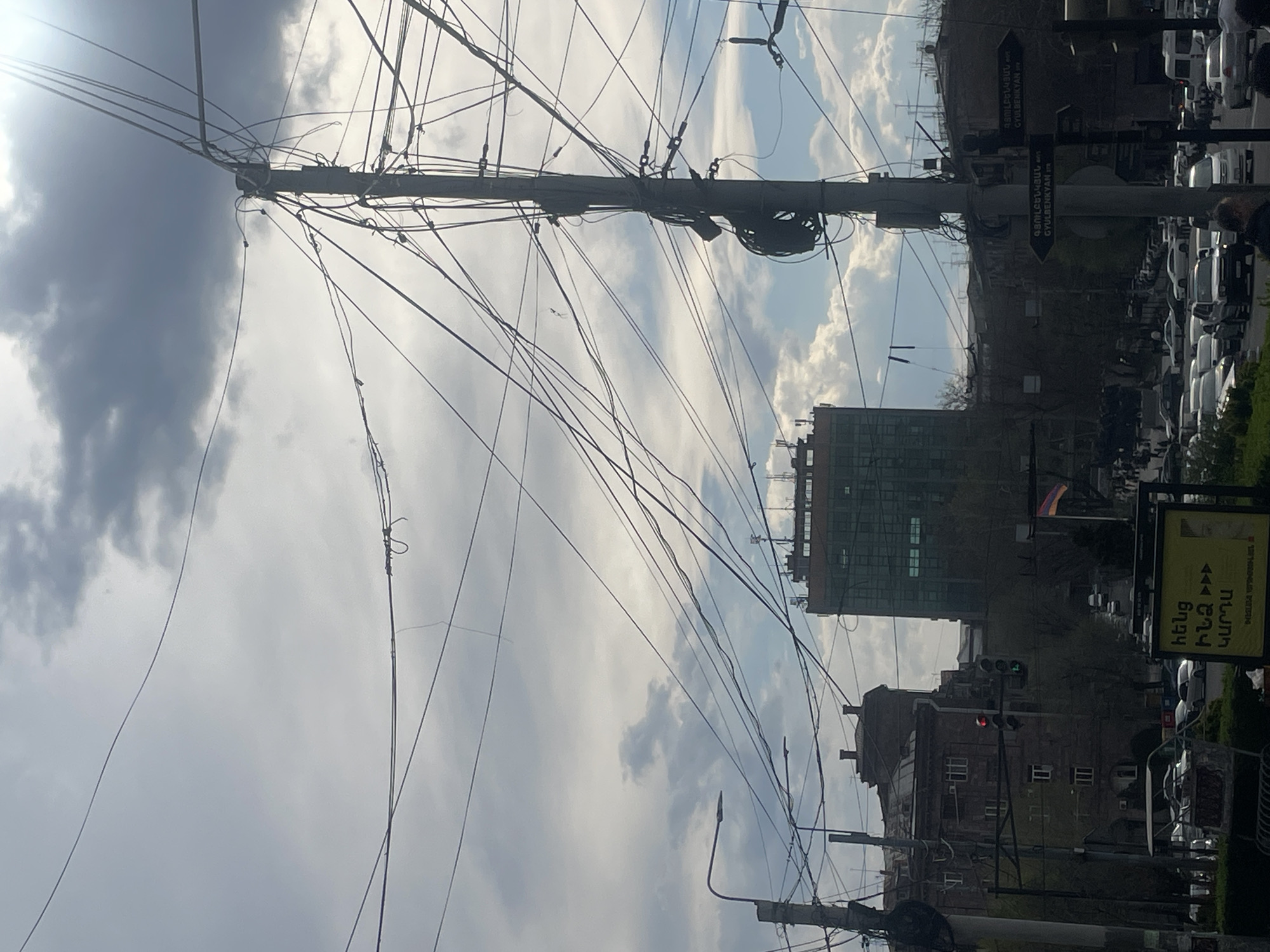 The image shows a street scene in Yerevan, Armenia, characterized by a dense network of overhead power lines obscuring the sky. The view is angled upwards towards a utility pole laden with cables. In the background, there are buildings, including a multi-story office or residential building with a modern glass facade, and an older building with a traditional architectural style. Street signs in Armenian script and a billboard are visible. The Armenian flag is seen, adding to the sense of location. Traffic is present along the street and people are passing by. The sky is overcast with some patches of sunlight.
