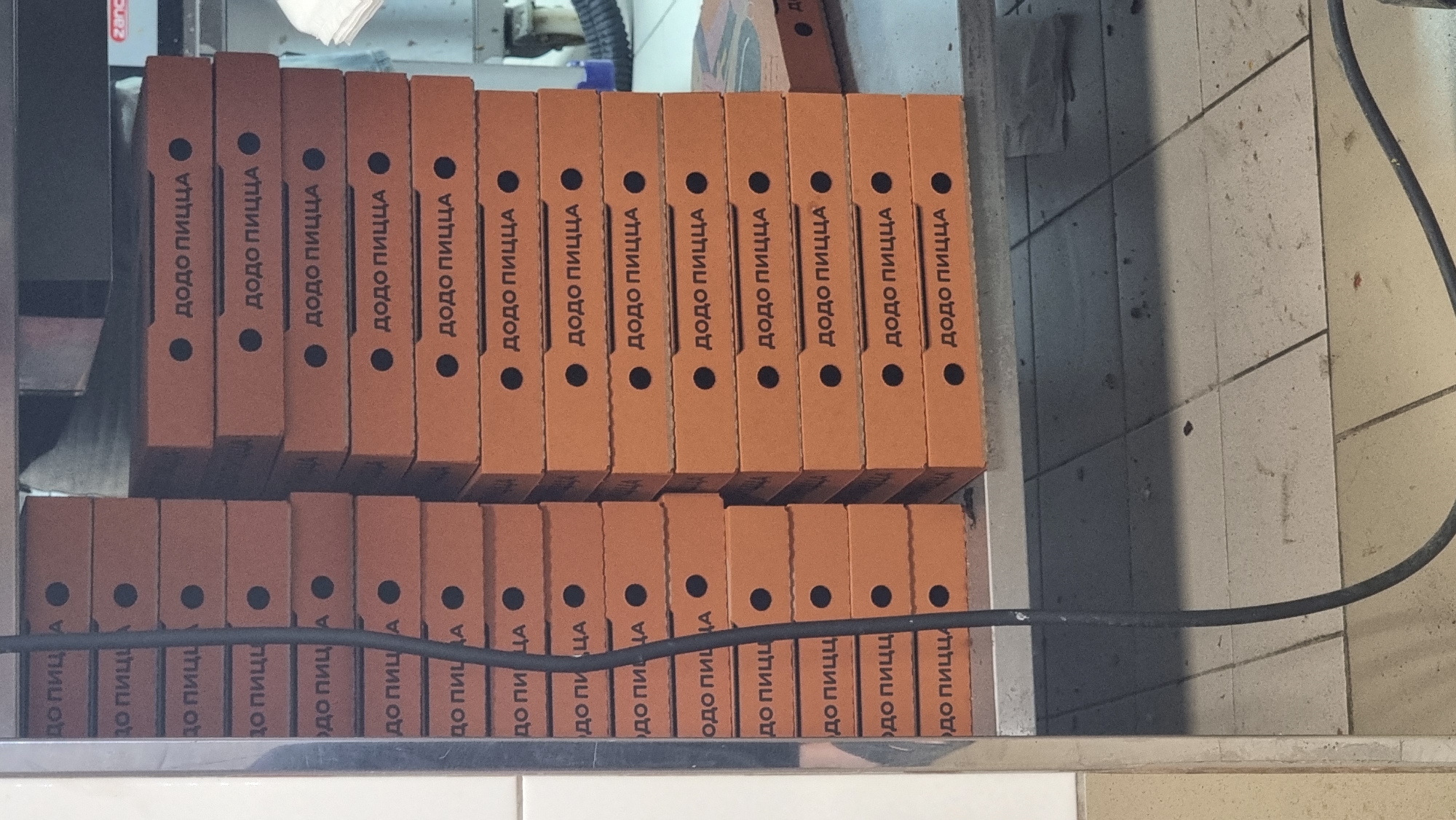 The image shows a stack of orange pizza boxes with the text "ДОДО ПИЦЦА" in Russian, meaning "Dodo Pizza," printed on them. The boxes are stacked in two columns, one to the left and one to the right, on a metal shelf. A black cable runs vertically down the center of the image, obscuring part of the left stack of boxes. The environment appears to be a storage or preparation area, possibly a kitchen or stockroom, with tiled flooring visible below the shelf. A small red and white logo, possibly a brand name, is visible on the far right of the shelf. No people are present in the image. The scene is a straightforward depiction of stacked pizza boxes.