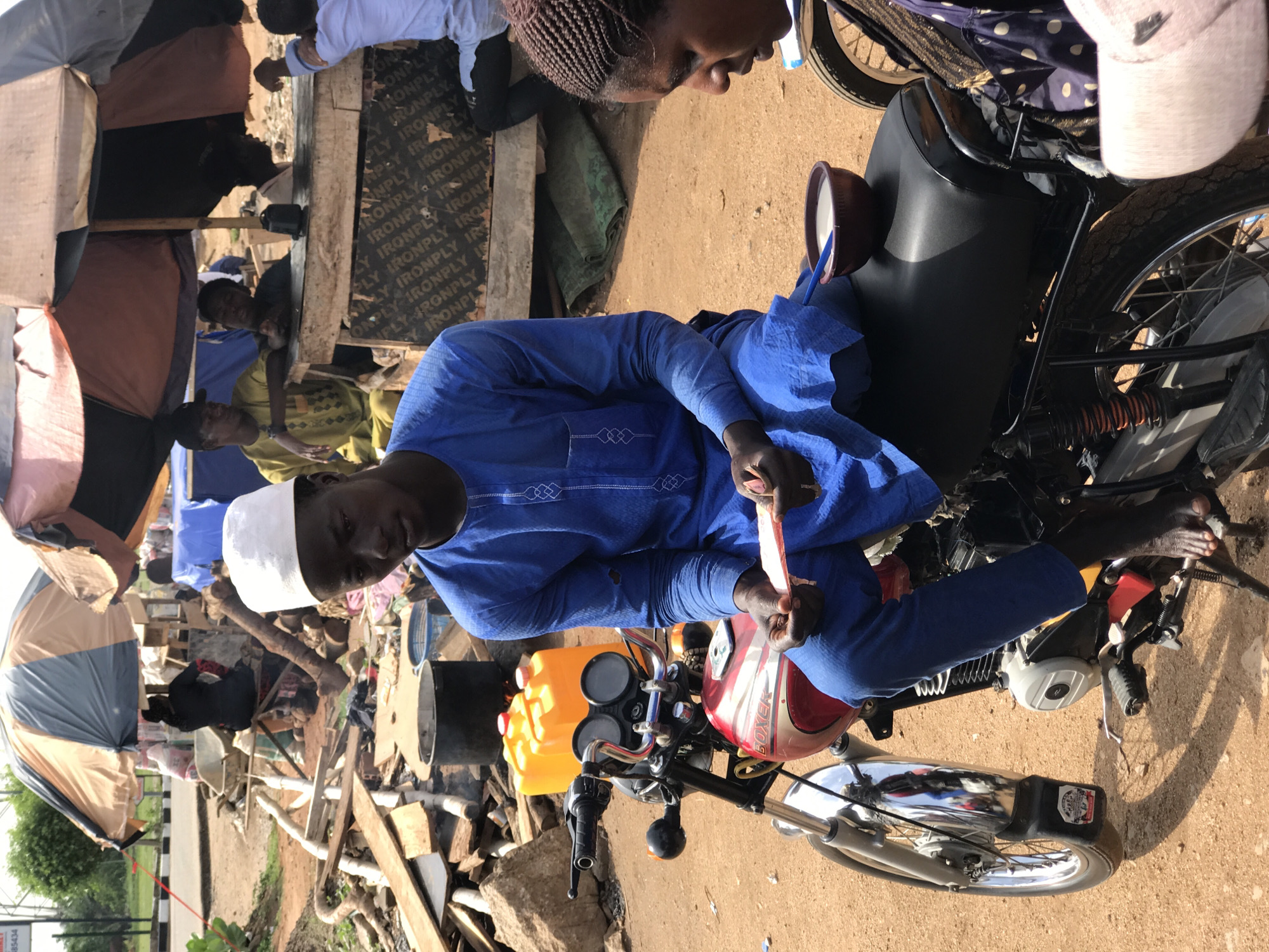 Here's a forensic description of the image:
**Scene:** An outdoor setting in Gwarinpa, Nigeria. The ground is dirt.
**People:** A man wearing a blue traditional outfit and a white kufi cap is seated on a motorcycle. His facial features are visible. Several other individuals are present in the background, exhibiting a range of traditional African attire.
**Objects:** A motorcycle with a yellow fuel tank and a red helmet placed on the handlebars is the central object. Debris, including pieces of wood and construction materials, are scattered around. There is also an umbrella standing nearby.
**Actions:** The man is seated on the motorcycle, holding an unidentified object in his hands. Other people in the scene are standing or sitting in the background.
**Interactions:** The man on the motorcycle is the main subject of the image, but he seems to be interacting with people in the background.
**Environment:** The overall environment suggests a somewhat informal or unplanned setting, with scattered debris indicating possible construction or recent activity.