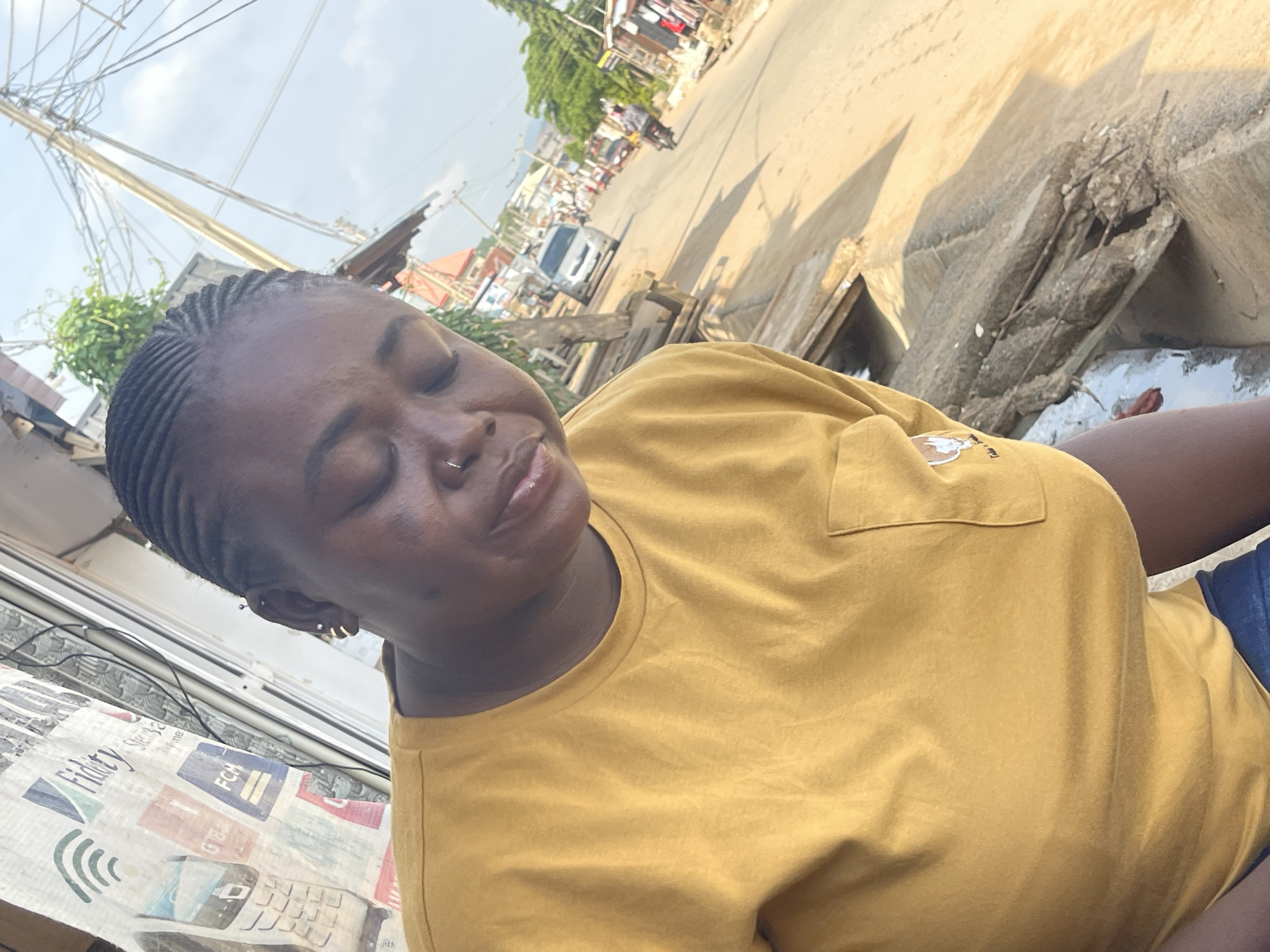A close-up, eye-level outdoor shot features a dark-skinned woman with braided hair and a nose ring. She is wearing a mustard yellow t-shirt with a small pocket on the left side of the chest, and a faint logo is visible on the pocket. Her eyes are closed and her head is tilted slightly down. The background reveals a street scene in Abuja, Nigeria. A busy road stretches into the distance, with vehicles including a silver minivan, motorcycles, and other cars. Buildings and market stalls line the sides of the road, and power lines are strung overhead. The ground appears to be a mix of dirt and concrete. The lighting suggests it is daytime, with bright sunlight casting shadows.
