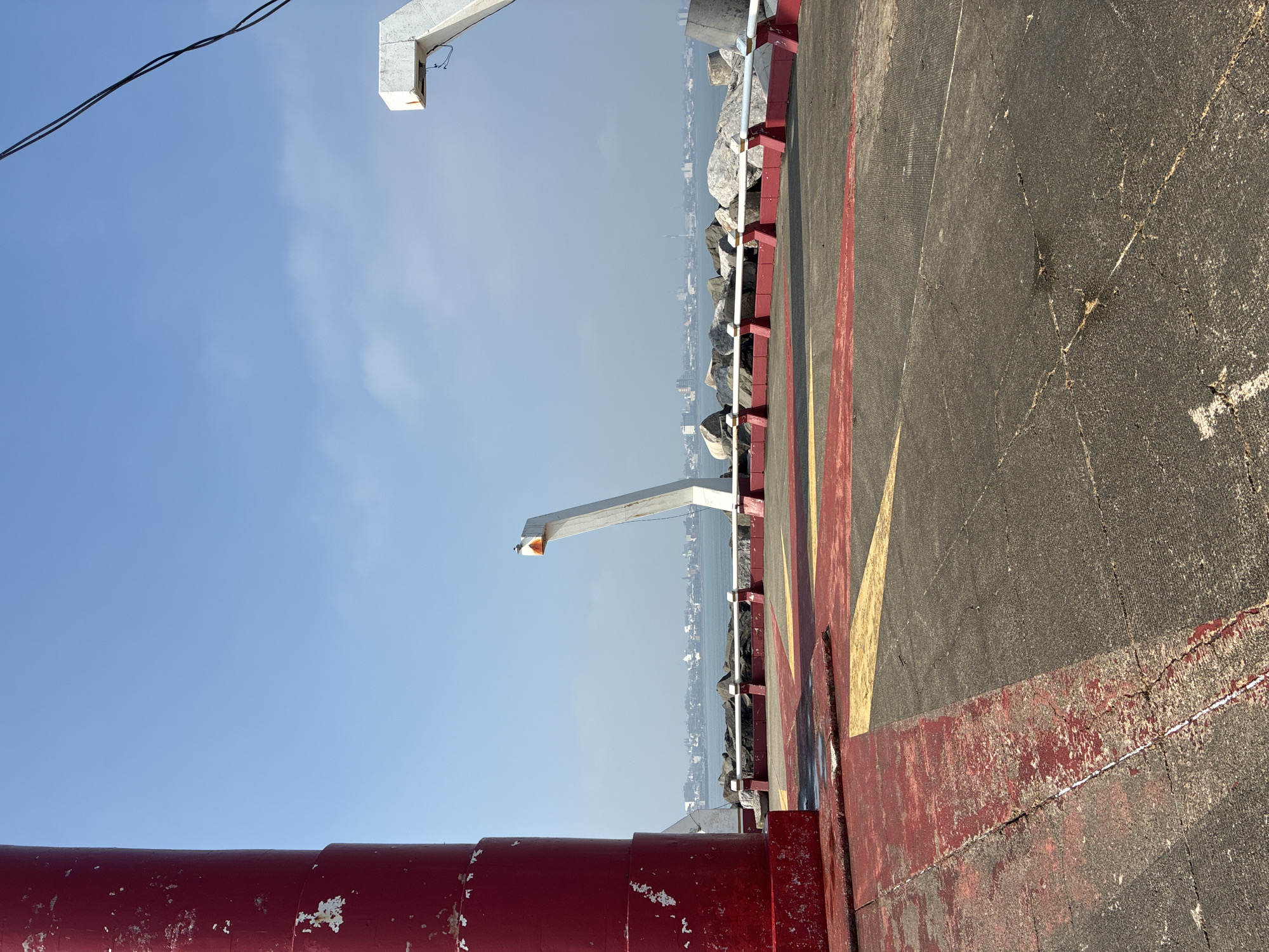 The image shows a concrete pier extending into the sea. Along the edge of the pier are large rocks, with a cityscape visible in the distance. On the right, the pier surface is marked with yellow and red stripes. A red railing runs along the foreground of the pier, and metal support structures are positioned above and at the far end. The sky is blue with some clouds. The location is likely in Coatzacoalcos, Mexico, given the coastal setting and urban landscape. There are no people visible in the scene.