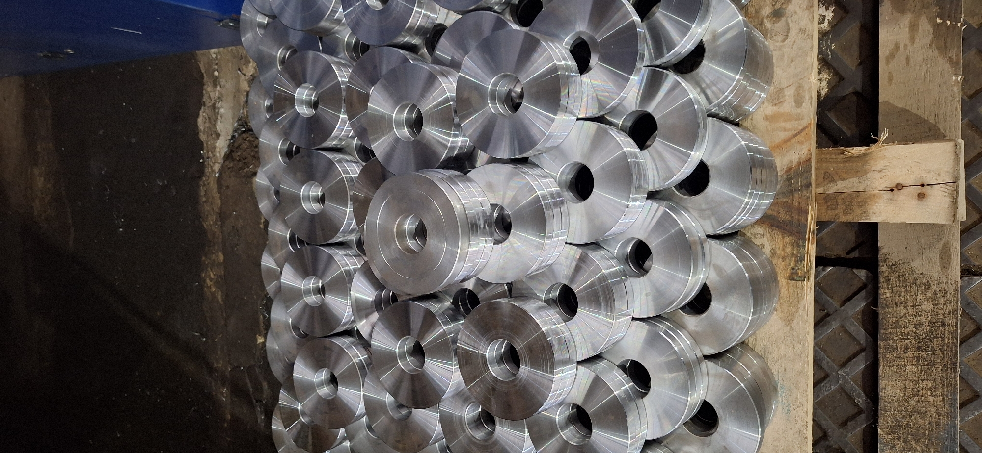The image shows a large pile of identical, circular metal components with a hole in the center, stacked on a wooden pallet. The components appear to be machined and have a metallic, silver finish with visible machining marks. The background is dark and appears to be a workshop or industrial setting, with a portion of a blue machine visible on the right side. The scene is devoid of people. The location is stated to be Nakhodka, Russia, implying an industrial or manufacturing context within that city.