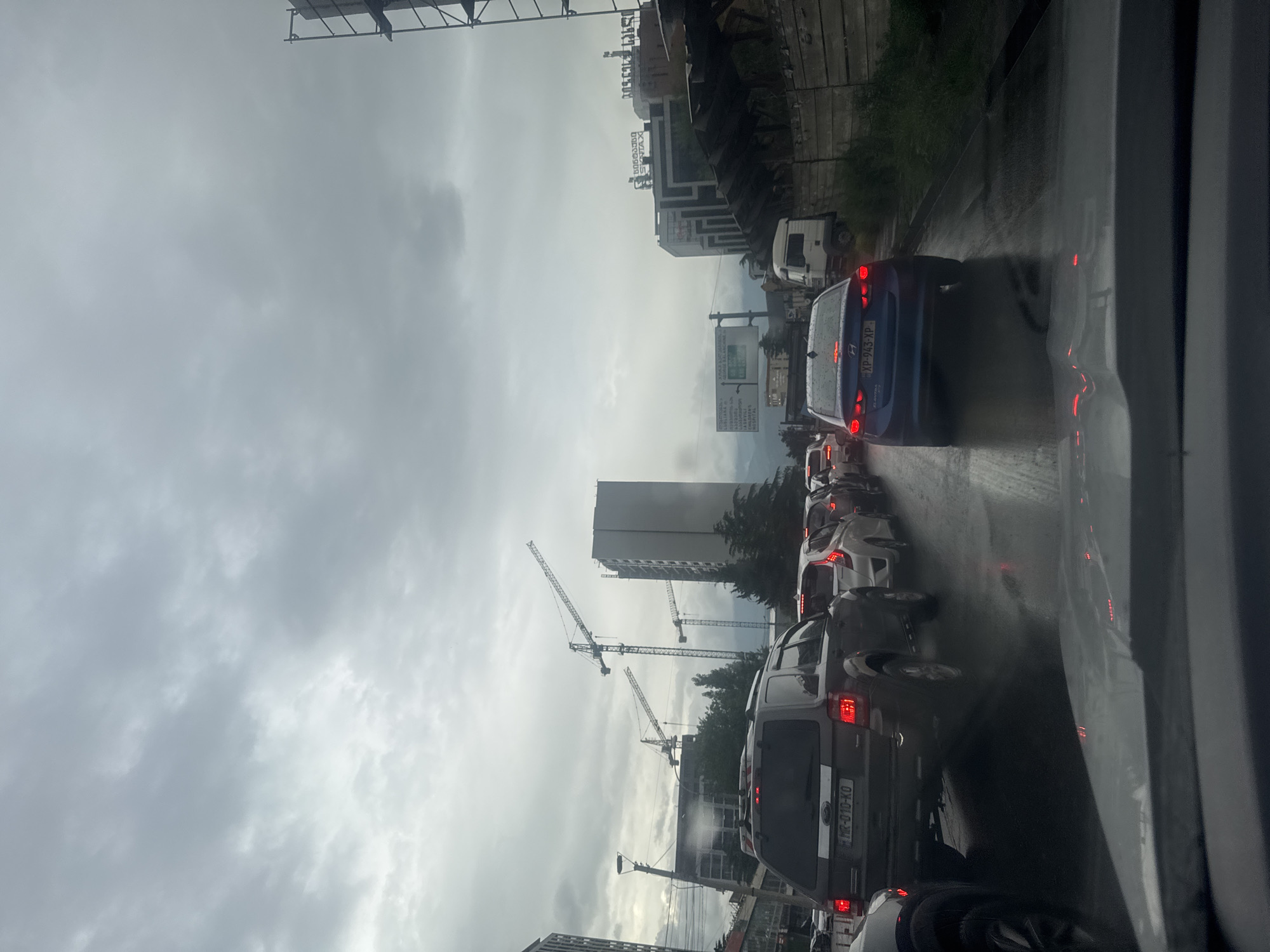 The image depicts a traffic jam on a rainy day in Tbilisi, Georgia. A line of vehicles stretches along a wet asphalt road. Several construction cranes are visible in the background, alongside a tall building under construction. Other buildings and a billboard are also present in the scene. The sky is overcast with gray clouds. The photograph was taken from inside a vehicle.
