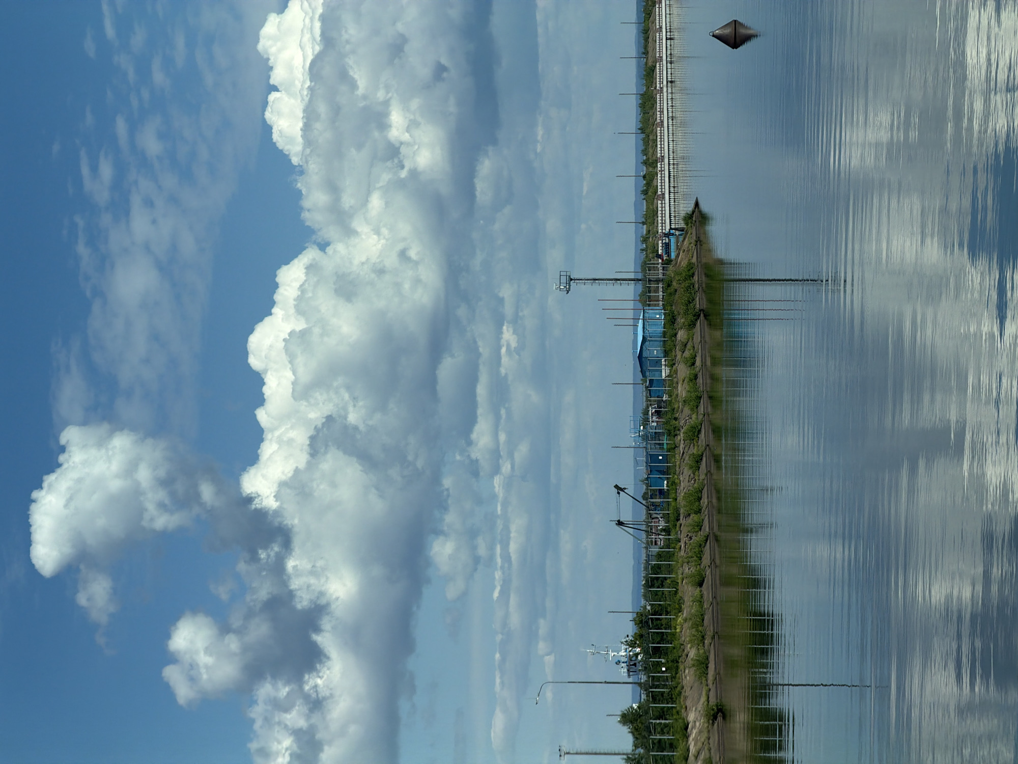 This detailed image depicts a calm industrial waterscape in Chaikovsky, Russia, under a dynamic sky. The upper half of the frame is dominated by a bright blue sky filled with a multitude of large, volumetric white and grey cumulus clouds, casting soft shadows and reflecting on the water below.
The lower half of the image features a still body of water, exhibiting subtle ripples that create textured reflections of the sky, clouds, and shoreline structures. The water appears deep blue-grey.
Along the left and mid-ground shoreline, a concrete embankment supports a strip of green vegetation, including grass and small bushes. Above this, a metal fence with white vertical posts and a dark horizontal rail stretches along the bank. Visible on the shore are several structures: a light blue, rectangular building with a darker roof; a tall, dark lattice tower equipped with a platform and what appears to be sensing equipment; and a street light pole with a luminaire. A white and blue vessel, possibly a tugboat or workboat with a prominent mast, is partially visible behind the fence and vegetation on the far left. Further along the shore, a long, white, multi-segmented pier or dock extends parallel to the water, supported by numerous vertical posts. Behind the fence, various industrial-looking structures, including what appear to be cranes, are present.
In the far distance, a faint, low-lying landmass defines the horizon line. A dark, conical buoy with a flat top is positioned on the right side of the water, its reflection clearly visible on the surface. No individuals or active movements are depicted in the scene.