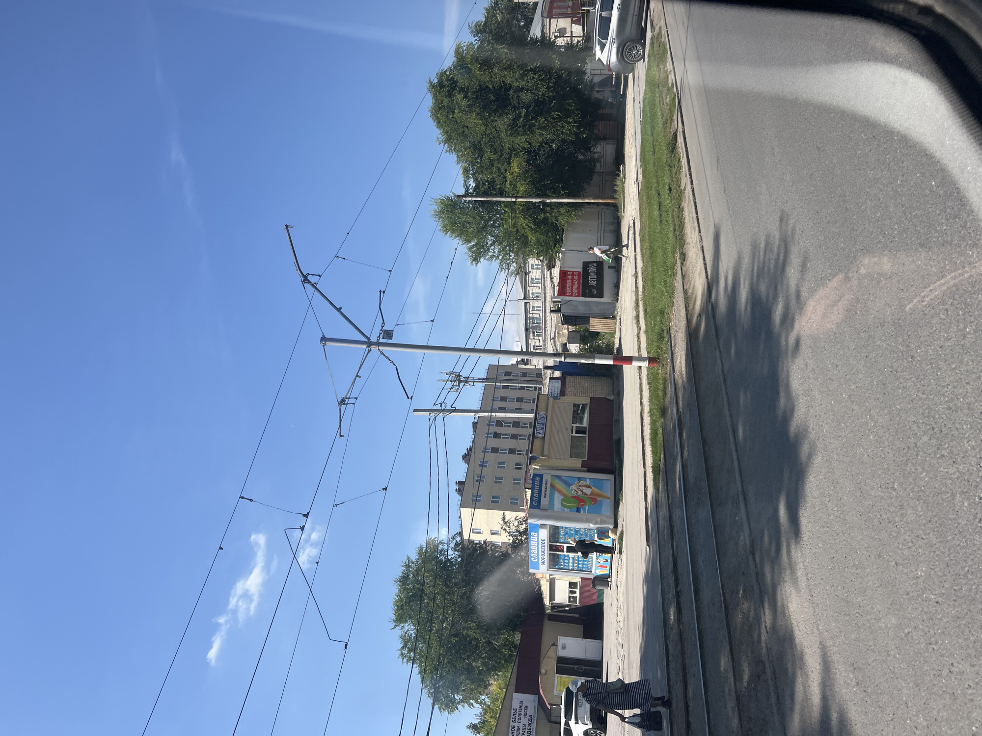 The image depicts a street scene in Ulyanovsk, Russia, on a clear, sunny day. In the foreground, tram tracks run along a paved road, with shadows of trees cast on the asphalt. To the left, a woman in a striped dress carries shopping bags. Adjacent to the tram tracks is a small kiosk labeled "СЛАВИЦА МОРОЖЕНОЕ" (Slavitsya Ice Cream), with an advertisement showing a colorful ice cream cone. Behind the kiosk are several low-rise buildings and a taller, multi-story building in the distance. Several trees with lush green foliage are visible, and overhead, numerous electrical wires for the tram system crisscross the sky. To the right, another kiosk or building is visible, and further down the street, a silver car is parked. Another person is seen walking away from the ice cream kiosk towards the right side of the frame. The overall atmosphere is that of a typical urban environment on a summer day.