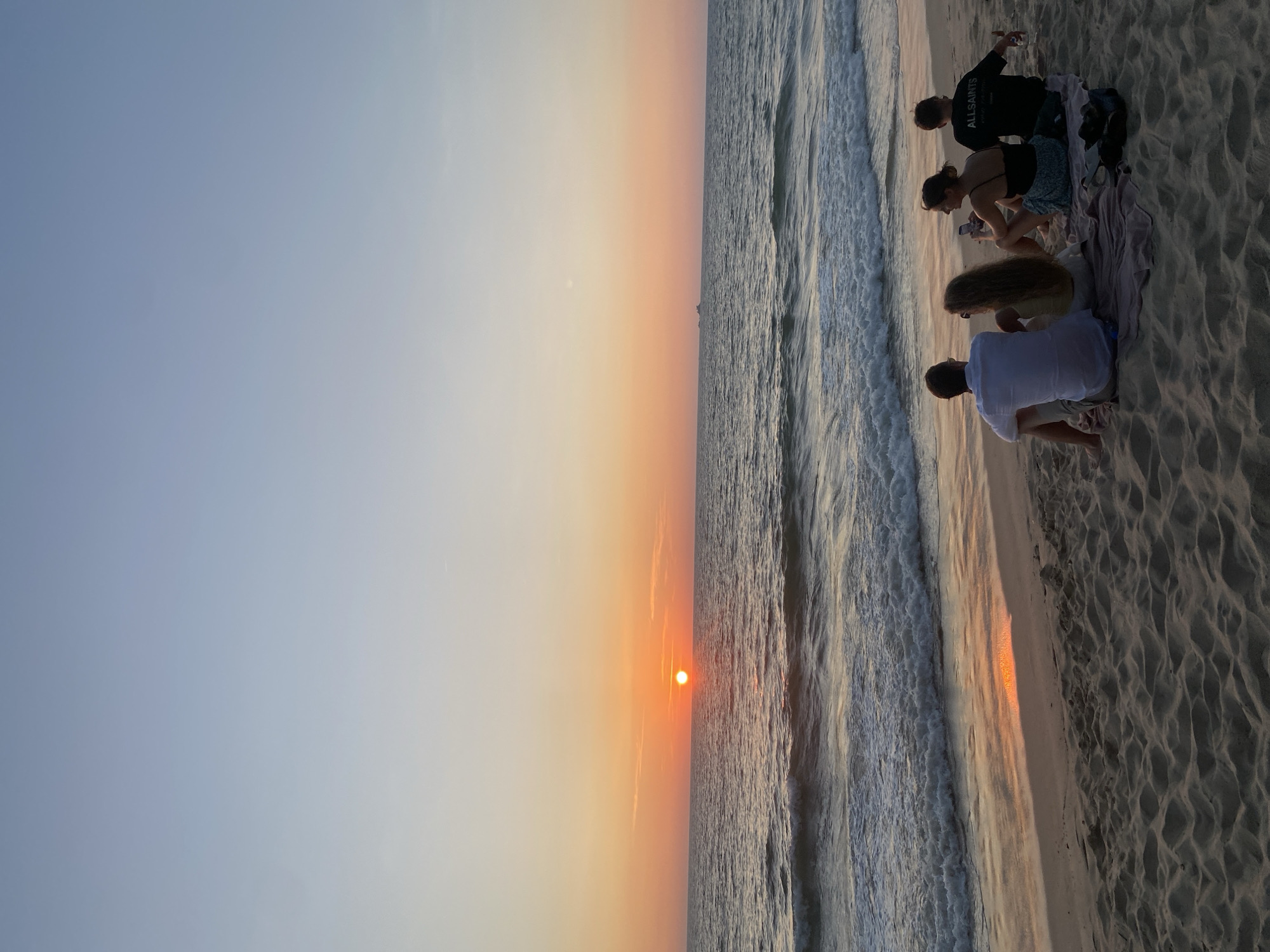 At a beach in Choeng Thale, Thailand, a group of four people are seated on the sand, observing the sunset. The sun is a bright orange disk setting over the ocean, creating a gradient of orange and yellow in the sky. The sea reflects the sunlight, with waves breaking on the shore. The people are seated close together on a towel on the sand. One individual is wearing a black shirt with "ALLSAINTS" printed on it, another is wearing a white shirt. The sand is textured and appears damp near the water's edge. The photo is taken at an angle, with the horizon dividing the image into a sky/sea section and a beach section.
