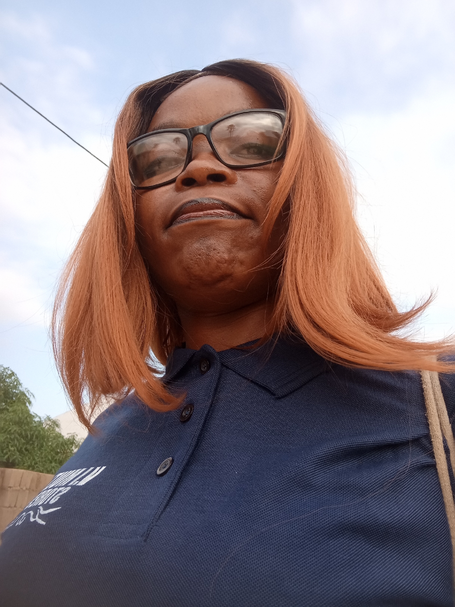 The image is a low-angle, close-up selfie of a woman wearing glasses and a dark blue polo shirt. She has reddish-brown hair and is looking down at the camera. The background appears to be an outdoor setting with a light blue sky and some foliage visible. A thin wire runs diagonally across the upper left portion of the image. There is a faint logo on the left side of her shirt. The photo was taken in the city of Kason, Nigeria.