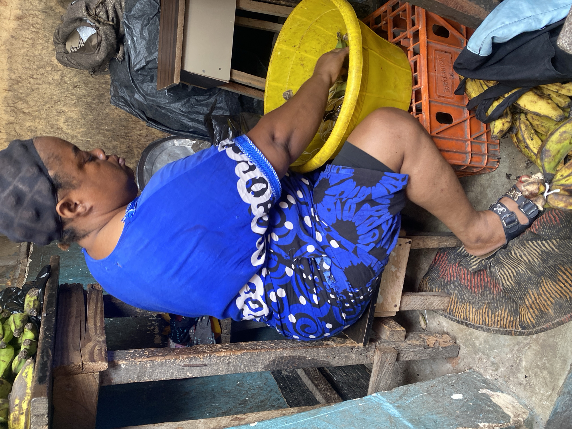 This image, taken in Ado, Nigeria, depicts a person, likely a woman, engaged in what appears to be market activity. She is shown from a side perspective, sitting or squatting on a rustic wooden structure composed of planks. She is wearing a bright royal blue shirt with white and black patterned sleeves and hem, along with dark shorts featuring blue and black floral patterns. A dark head covering completes her attire.
The central action shows her right arm reaching into a large, yellow plastic bucket. Inside and around the bucket are numerous bunches of green and yellowing bananas or plantains, suggesting she is sorting or arranging them for sale. More bananas are visible on the ground to her right.
The scene is set in what appears to be an informal outdoor or semi-sheltered market stall. Notable objects in the surroundings include an orange plastic crate, possibly for beverages, behind the yellow bucket, with "Schweppes" (or similar text) partially visible on its side. A light blue piece of fabric or clothing is draped over this crate. Other items include a black plastic bag near the back, a metal basin or bowl to her left, and a patterned fabric with red, orange, and black stripes on the ground near her foot, which is adorned with a black sandal featuring silver buckles and a butterfly design. The ground itself is a mix of concrete or packed earth with some faded blue paint. The environment suggests a utilitarian setting for commerce.