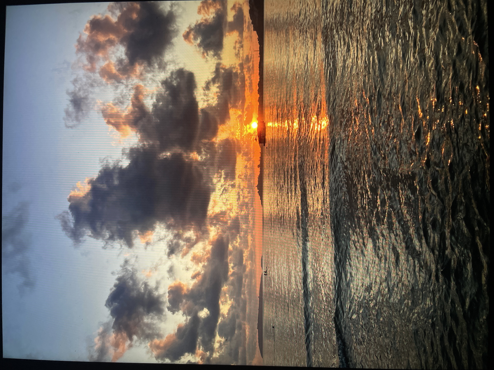 The image depicts a sunset scene at a beach. The sky is filled with clouds, some dark and heavy, tinged with orange and yellow from the setting sun. The sun itself is visible as a bright orb near the horizon. The ocean reflects the sunlight, creating a shimmering path across the water's surface. Two figures are visible on the beach in the distance. The city is unknown.